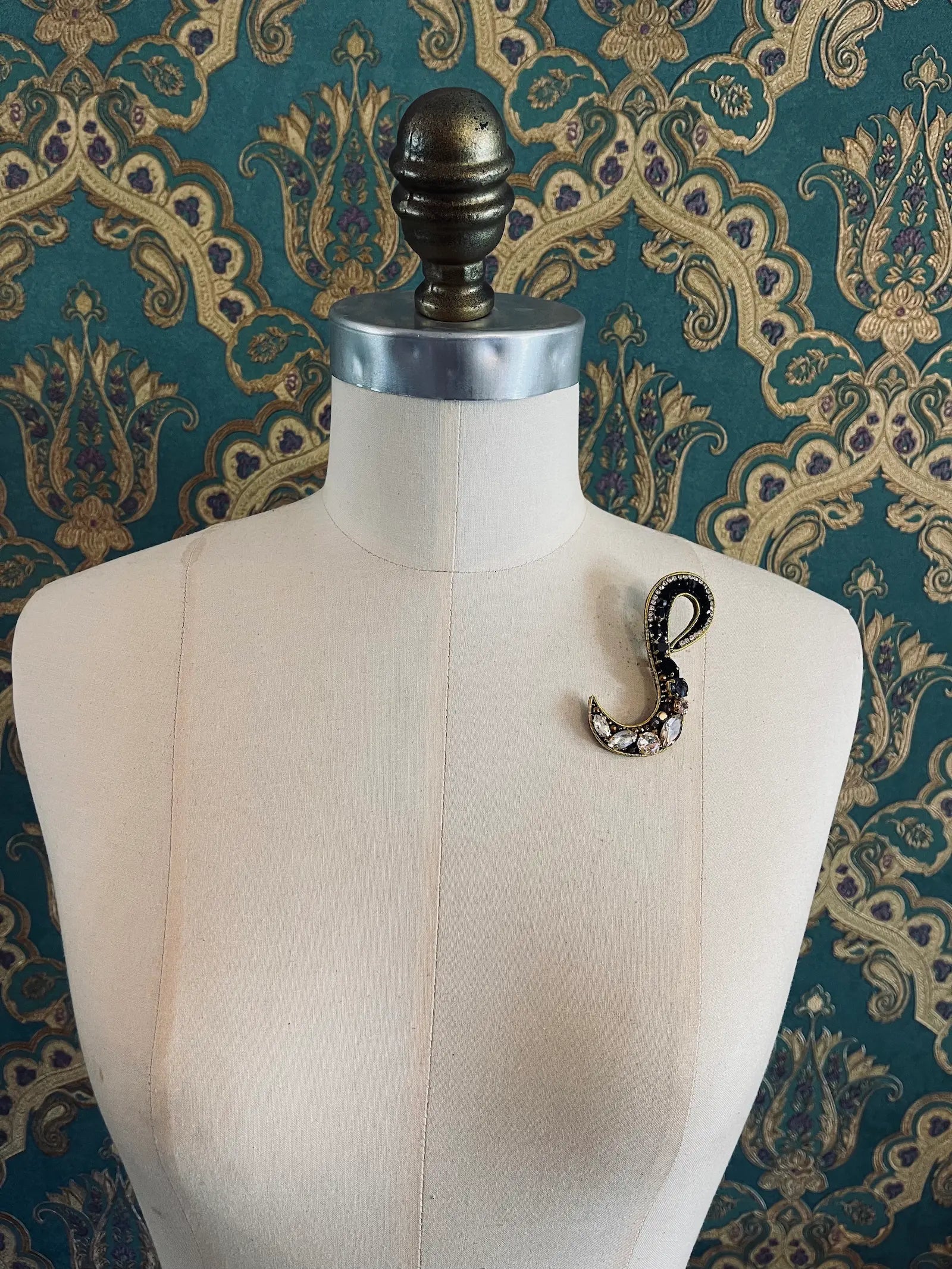 Broochella letter S hand beaded initial alphabet brooch in gold and black beads and sequins. Pictured on a mannequin. The background is green and gold damask wallpaper.