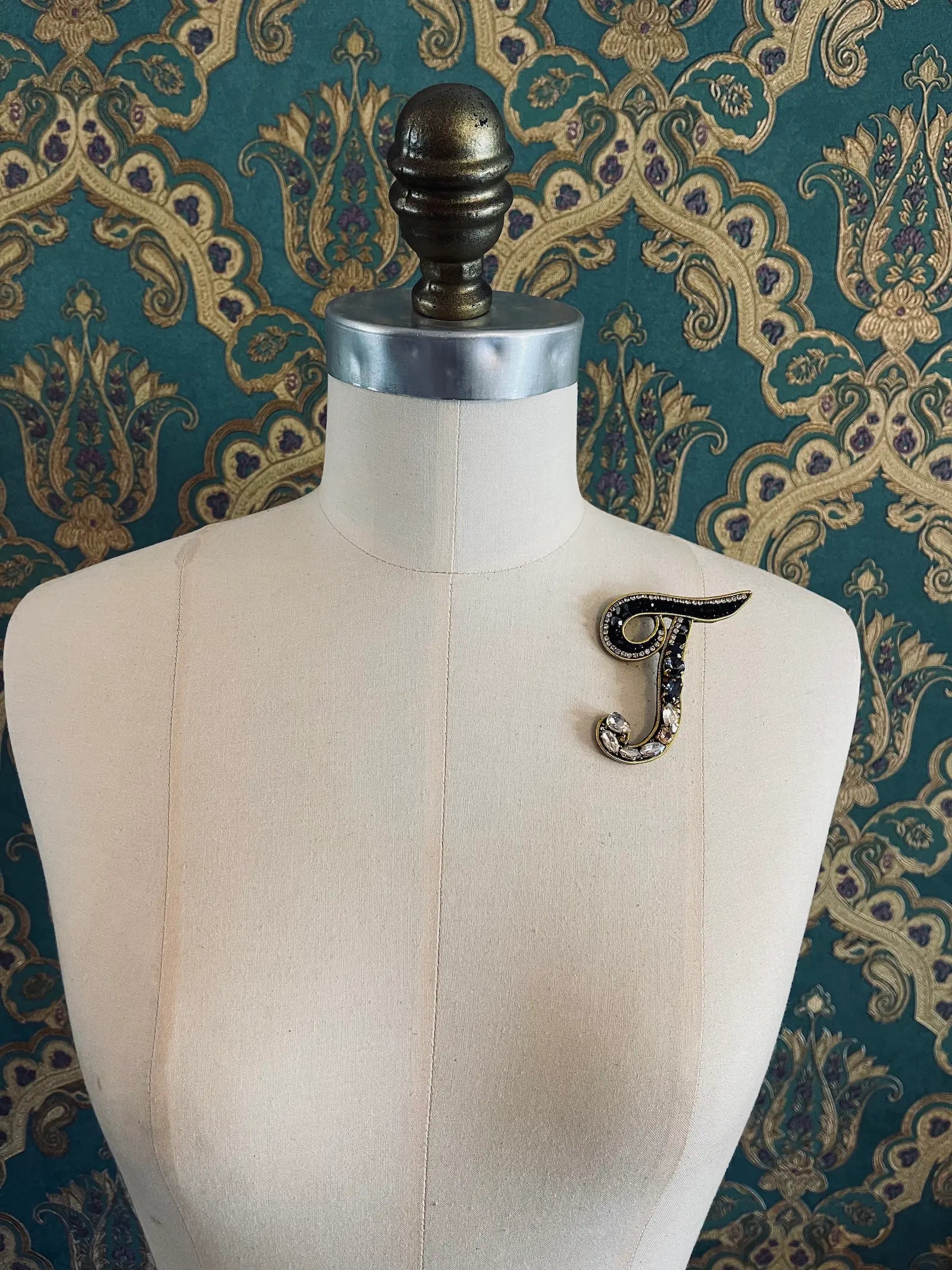 Broochella letter T hand beaded initial alphabet brooch in gold and black beads and sequins. Pictured on a mannequin. The background is green and gold damask wallpaper.