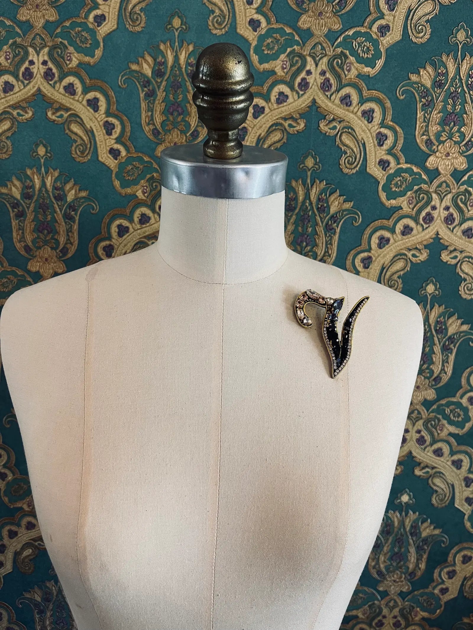 Broochella letter V hand beaded initial alphabet brooch in gold and black beads and sequins. Pictured on a mannequin. The background is green and gold damask wallpaper.