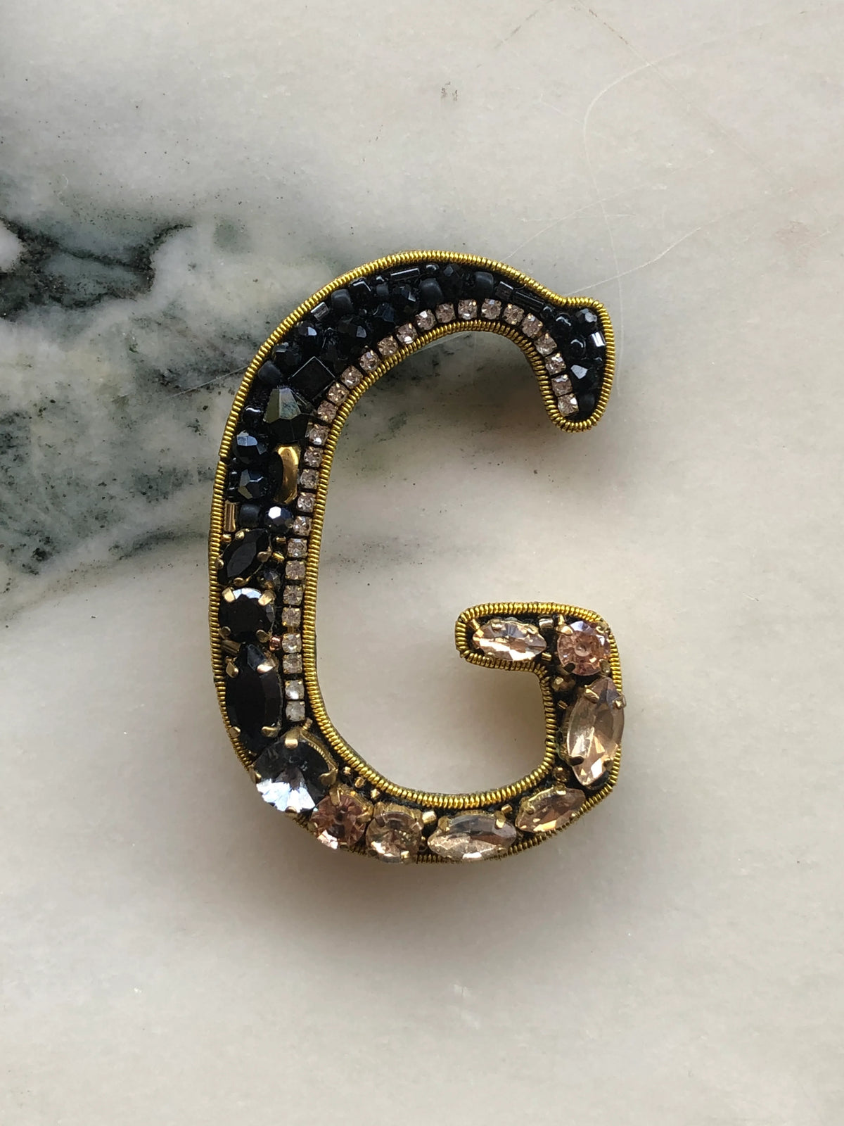 Broochella Initial G Brooch embellished with black beads, clear crystals and champagne stones with gold detailing, photographed on a white marble background with soft grey veining.