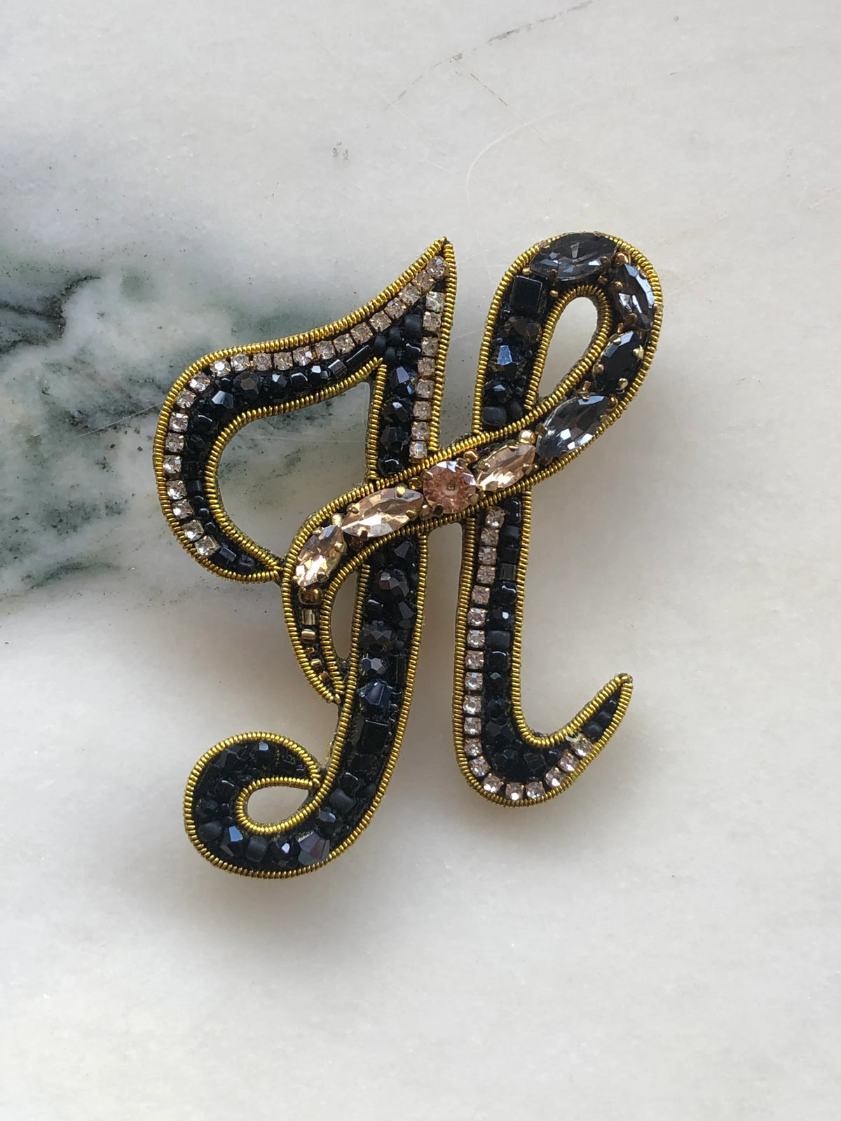 Broochella Initial H Brooch embellished with black beads, clear crystals and champagne stones with gold detailing, photographed on a white marble background with soft grey veining.