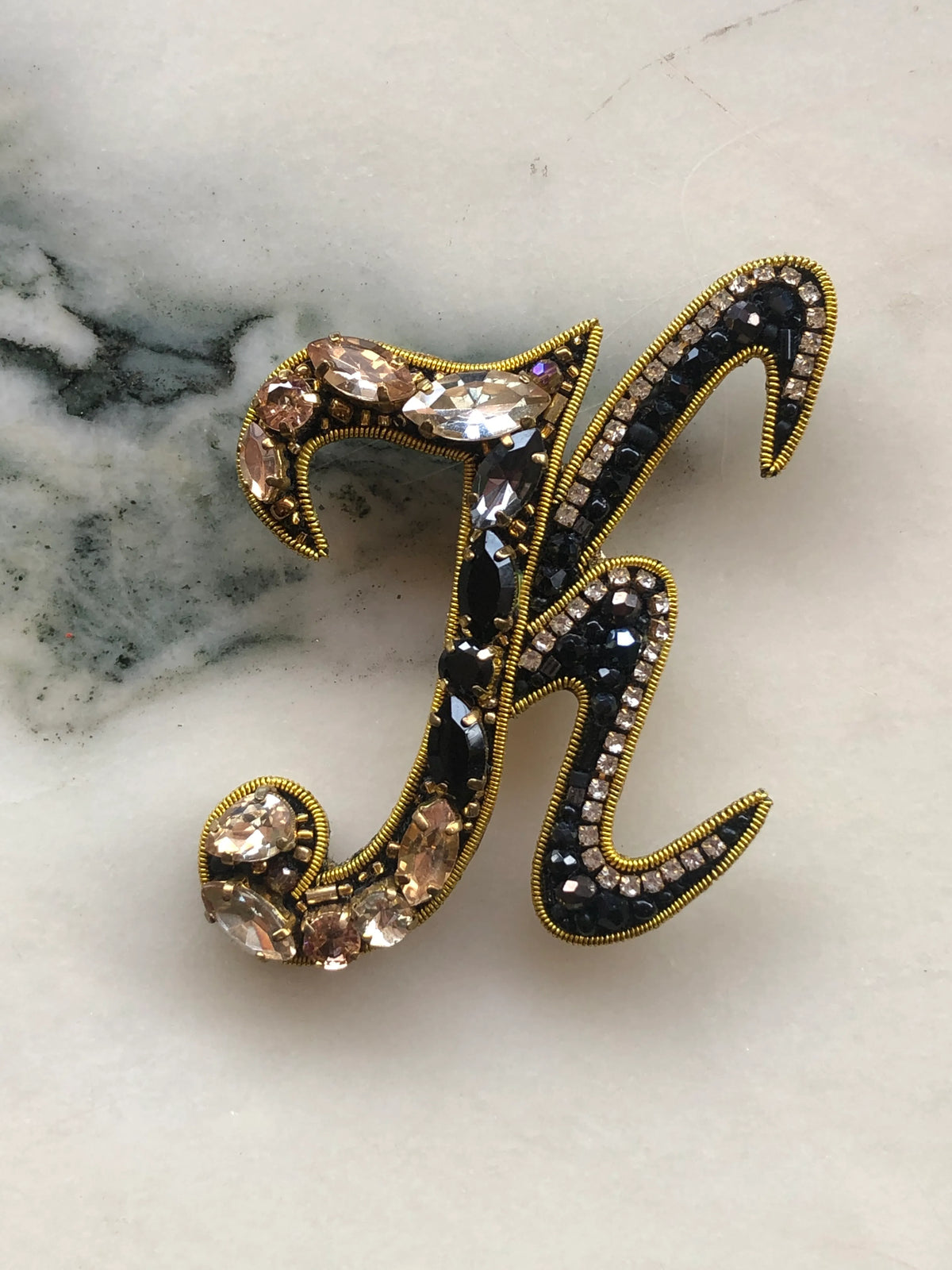 Broochella Initial K Brooch embellished with black beads, clear crystals and champagne stones with gold detailing, photographed on a white marble background with soft grey veining.