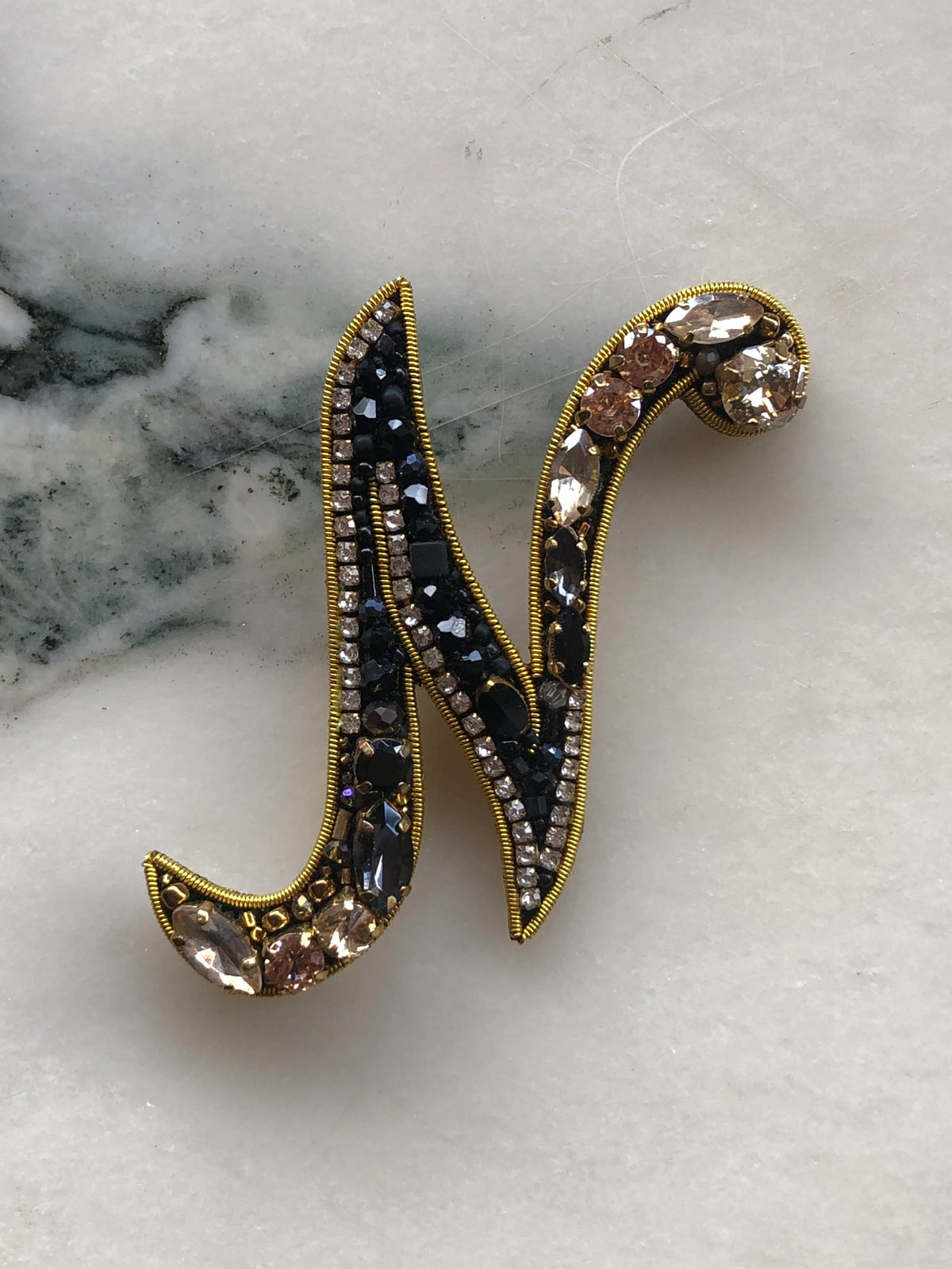 Broochella Initial N Brooch embellished with black beads, clear crystals and champagne stones with gold detailing, photographed on a white marble background with soft grey veining.