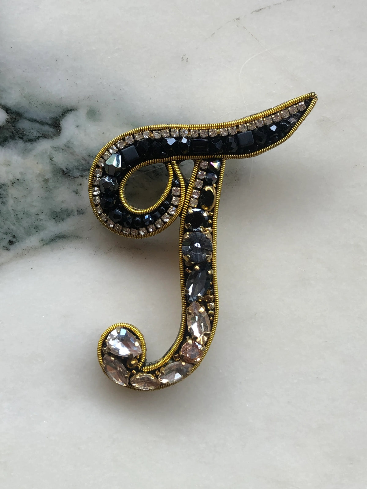 Broochella Initial T Brooch embellished with black beads, clear crystals and champagne stones with gold detailing, photographed on a white marble background with soft grey veining.