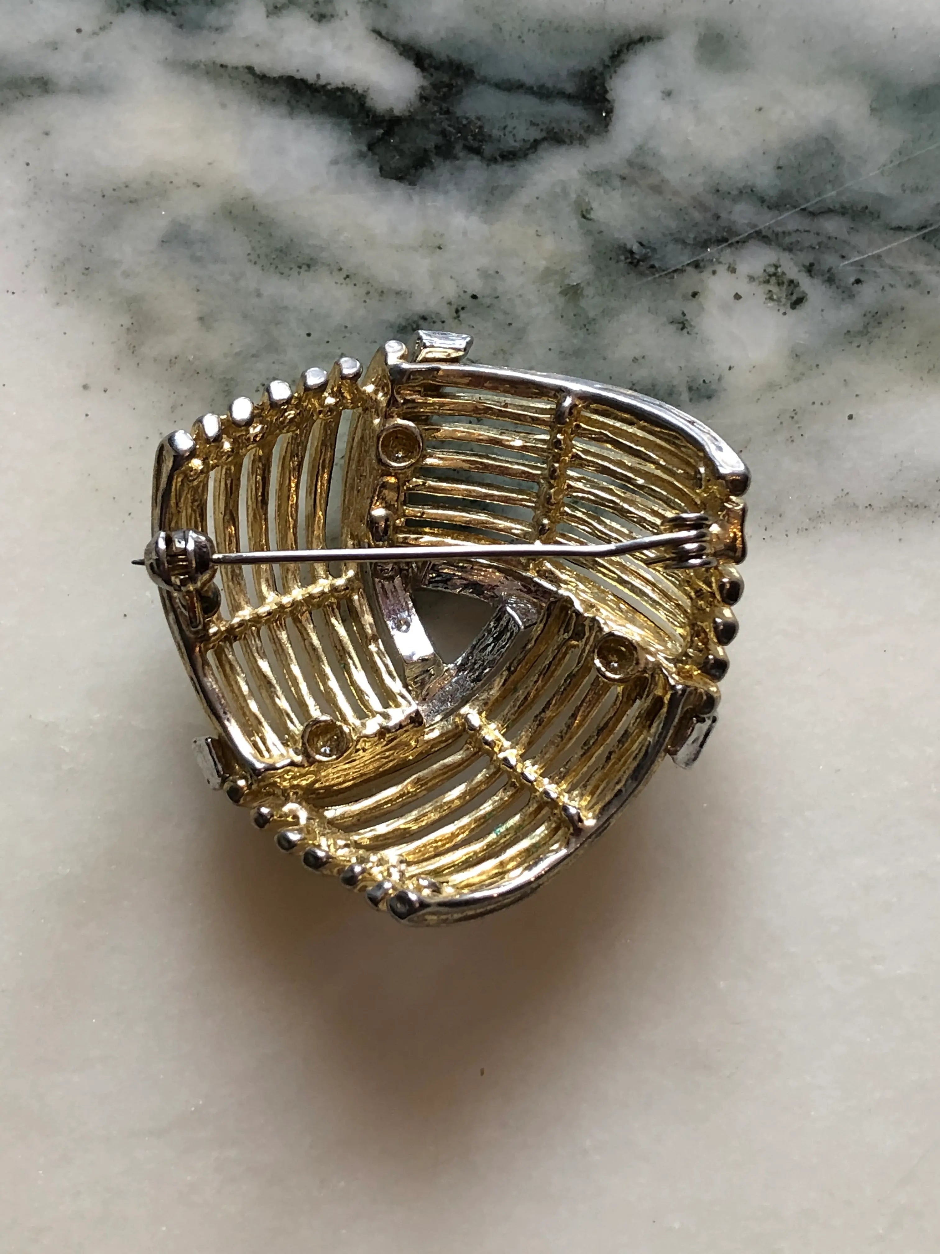 The back view of Broochella Intreccio vintage gold and silver-tone openwork brooch with sweeping ribbed bands and clear crystal accents. The background is white marble.