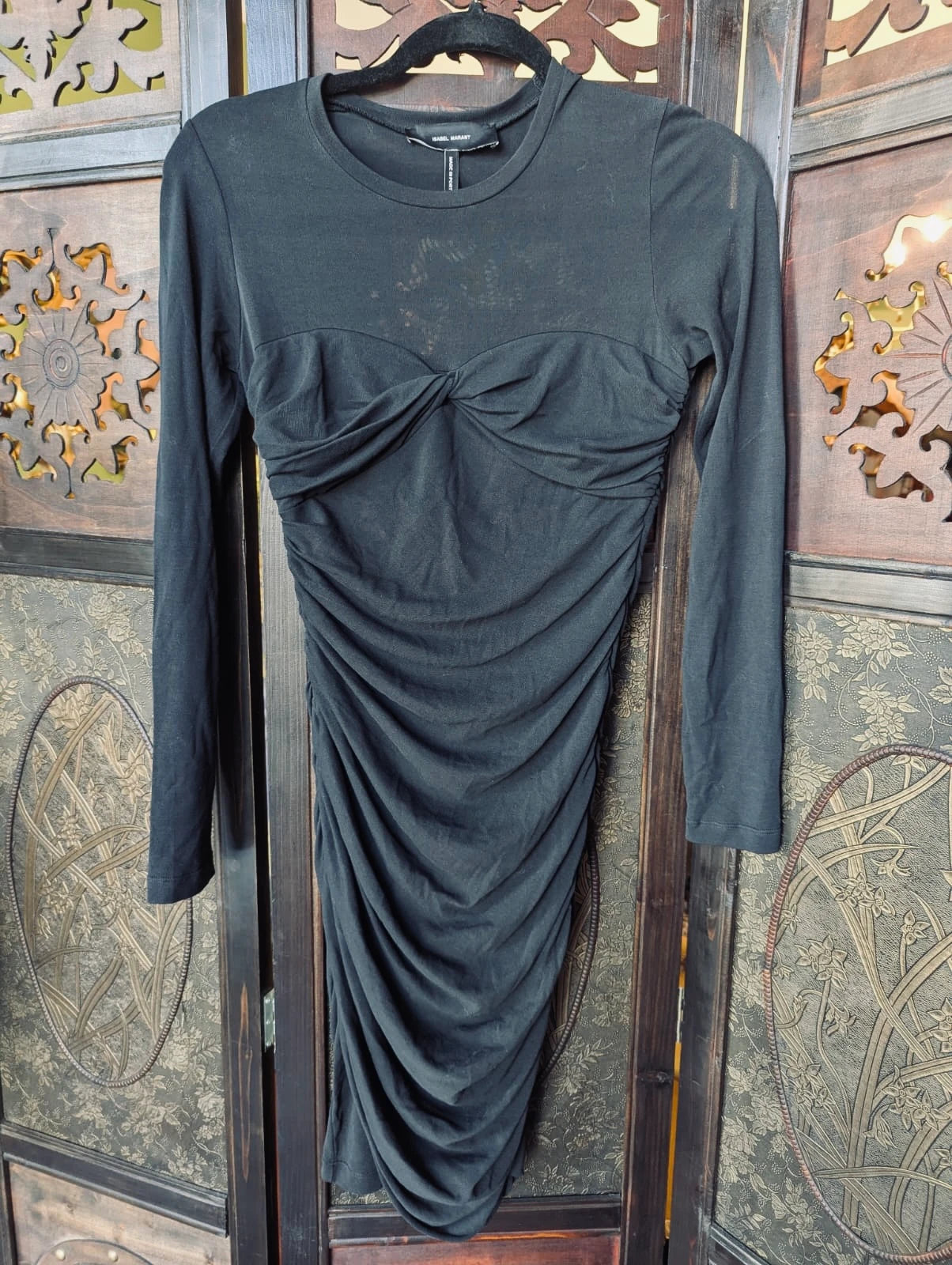 A Broochella  Isabel Marant Etoile black long-sleeve ruched dress with a sweetheart neckline and gathered bodice, hanging on a black hanger against a decorative wooden folding screen.