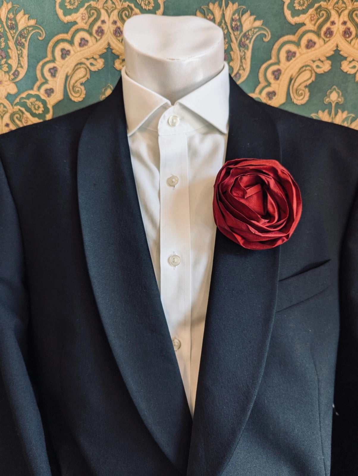 Broochella Isabella Rossellini handmade silk rosette flower brooch for men in burgundy. Pictured on a mannequin.  The background is green and gold damask wallpaper