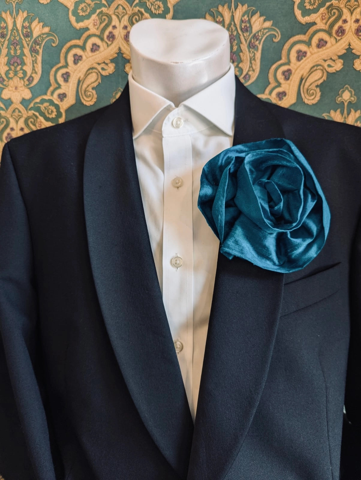 Broochella Isabella Rossellini handmade silk rosette flower brooch in peacock blue. Pictured on a mannequin.  The background is green and gold damask wallpaper