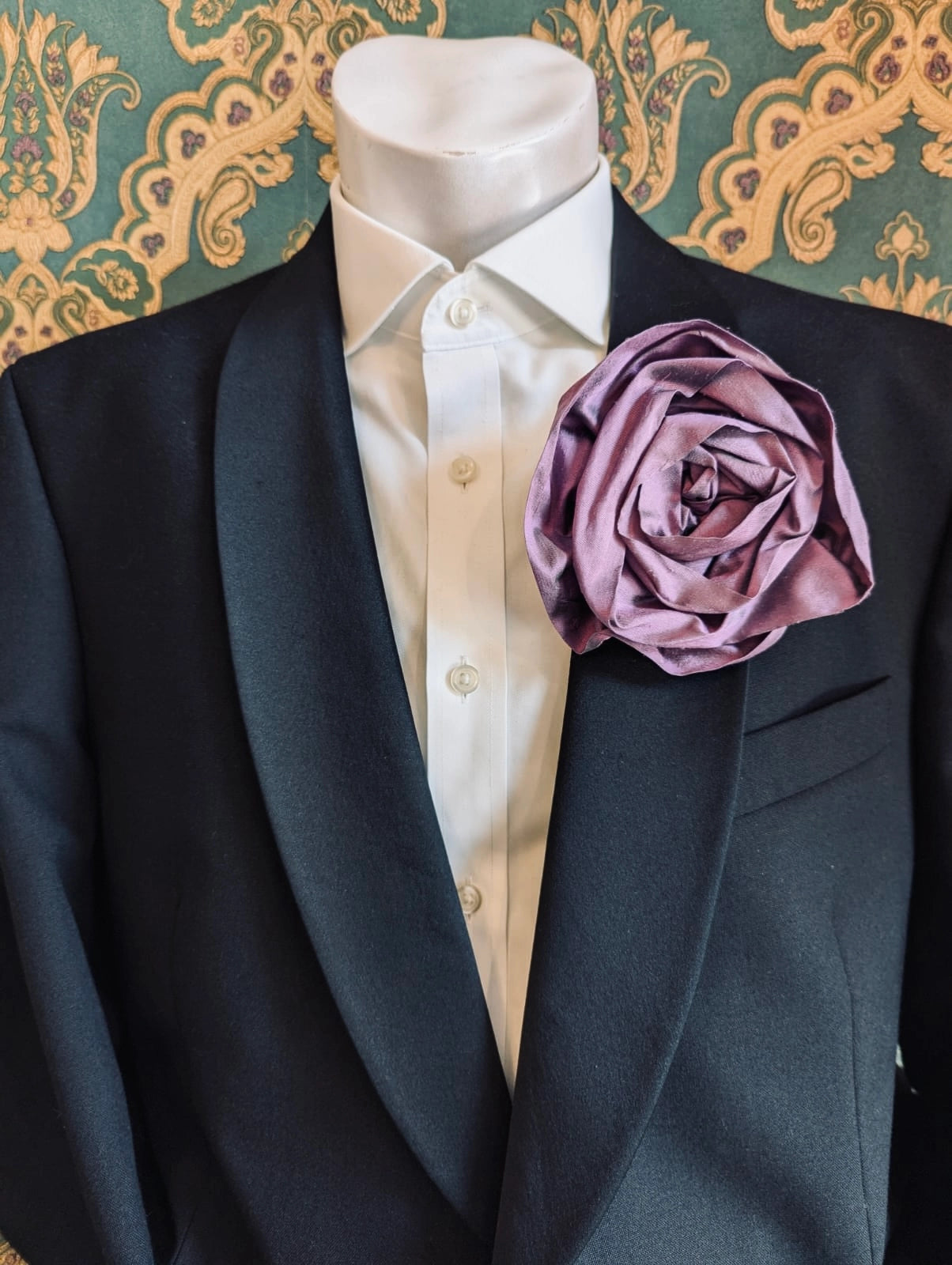 Broochella Isabella Rossellini handmade silk rosette flower brooch in violet. Pictured on a mannequin.  The background is green and gold damask wallpaper