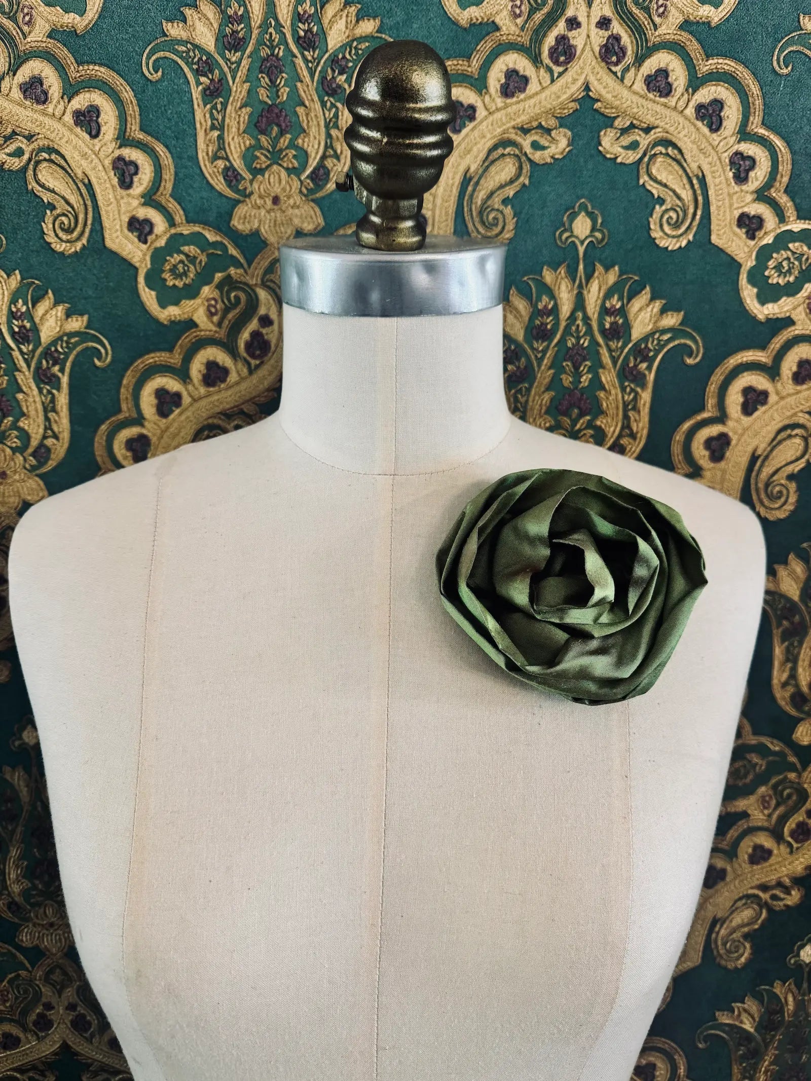 Broochella Isabella Rossellini handmade silk rosette flower brooch in green. Pictured on a mannequin.  The background is green and gold damask wallpaper