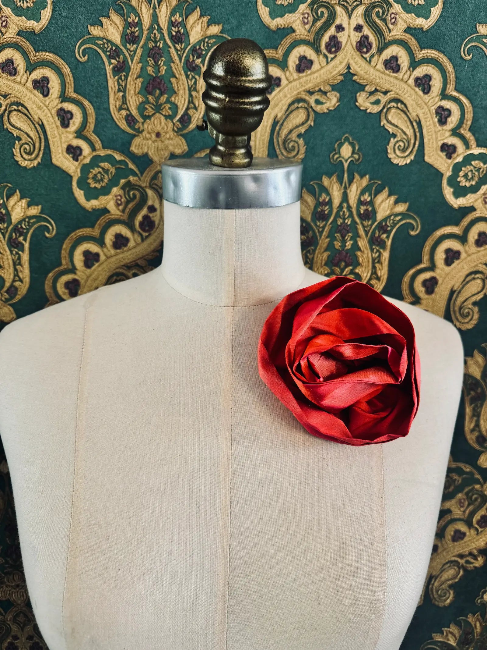 Broochella Isabella Rossellini handmade silk rosette flower brooch in tangerine. Pictured on a mannequin.  The background is green and gold damask wallpaper