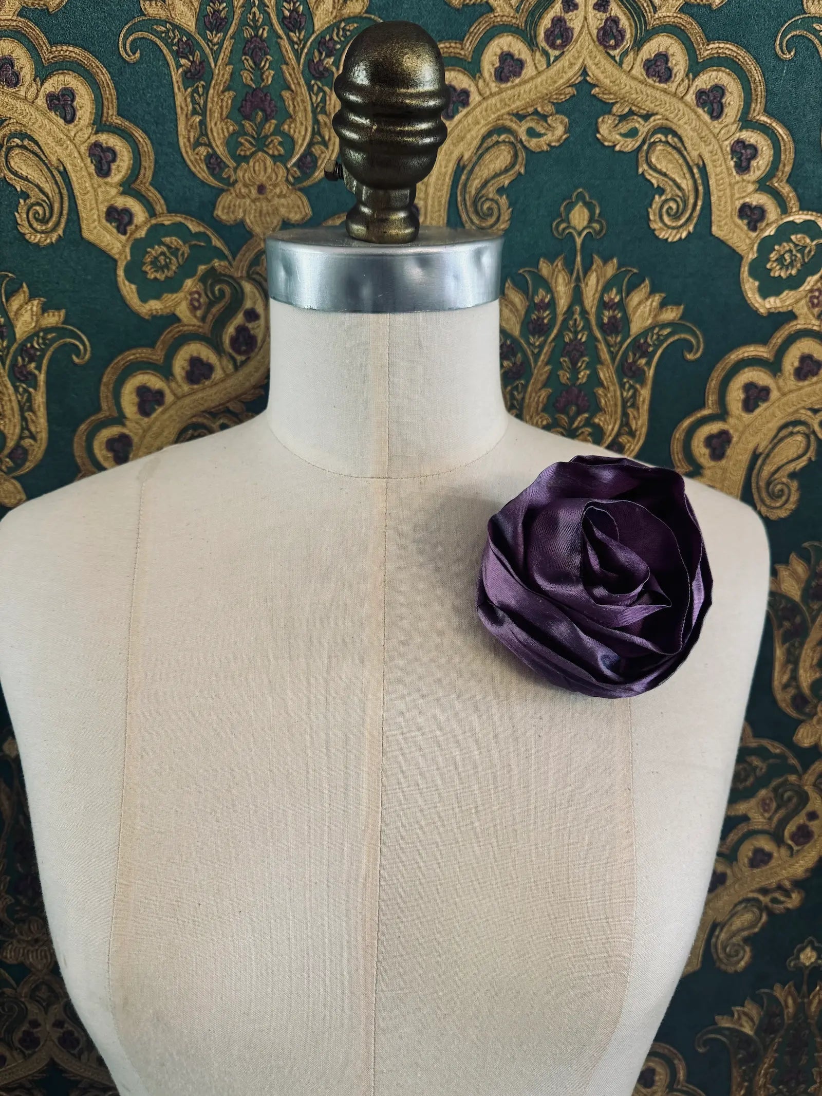 Broochella Isabella Rossellini handmade silk rosette flower brooch in violet. Pictured on a mannequin.  The background is green and gold damask wallpaper