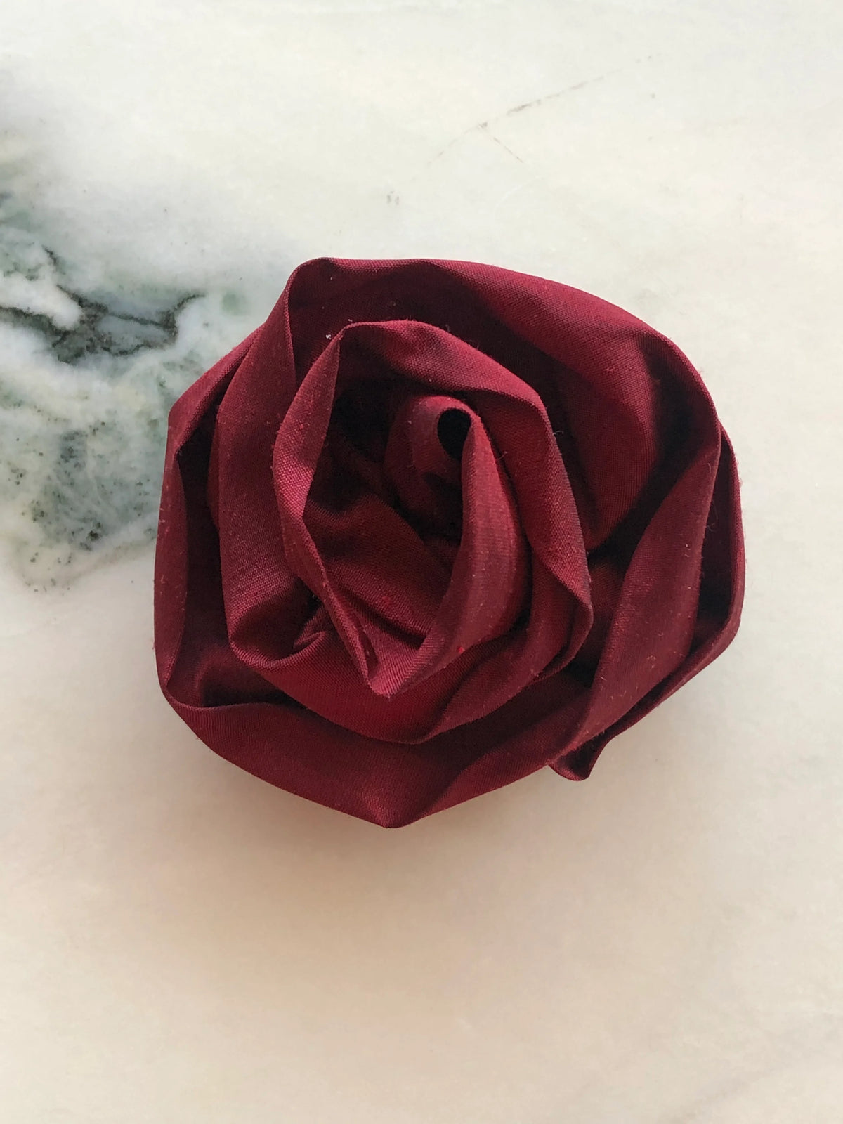 Handmade Broochella Isabella Rossellini designer silk rosette brooch for men in deep burgundy, a statement flower lapel accessory for suits and jackets, photographed on white marble.