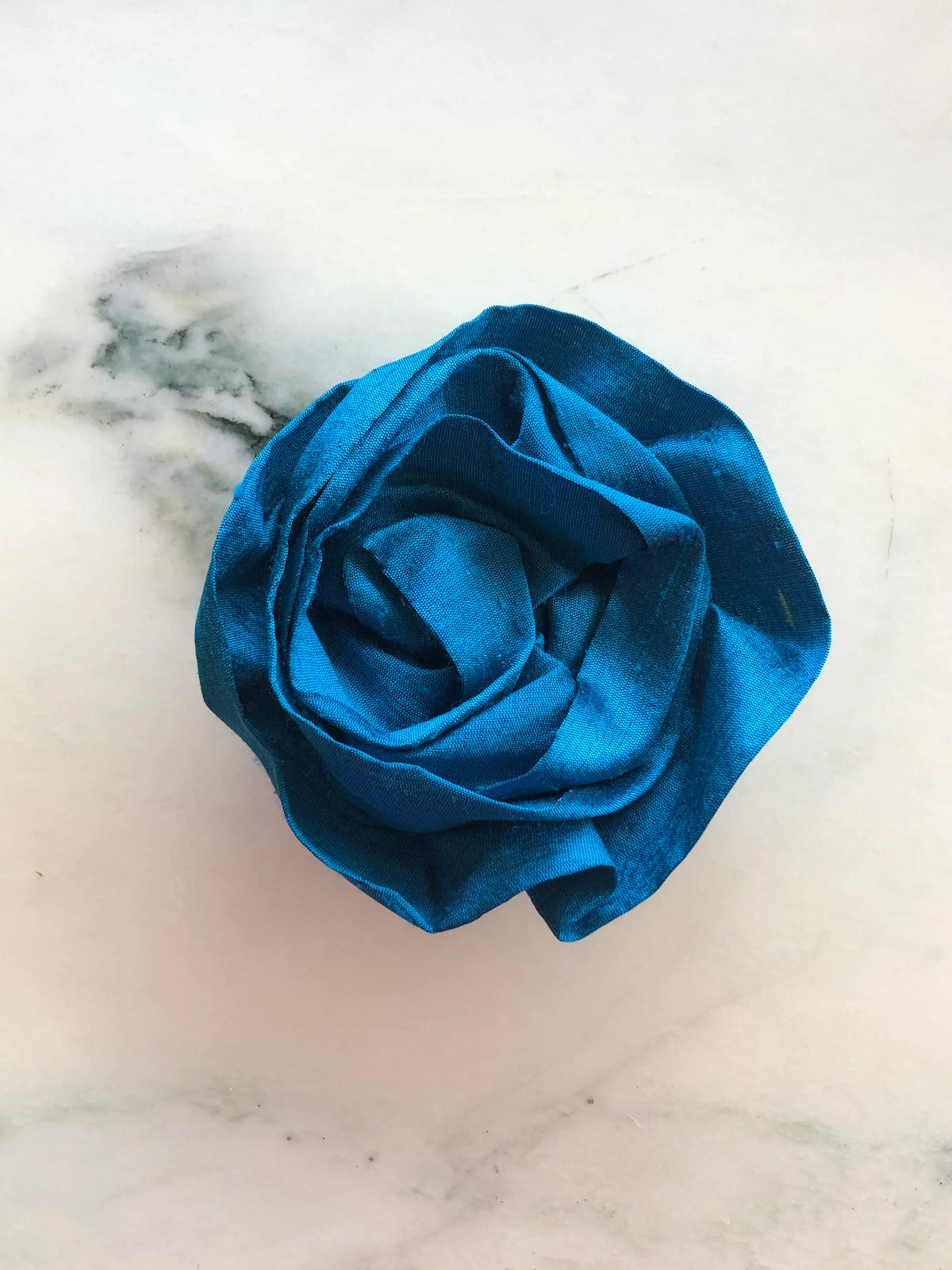 Handmade Broochella Isabella Rossellini silk rosette brooch in vibrant peacock blue, a designer statement flower with dimensional folded petals, photographed on white marble.