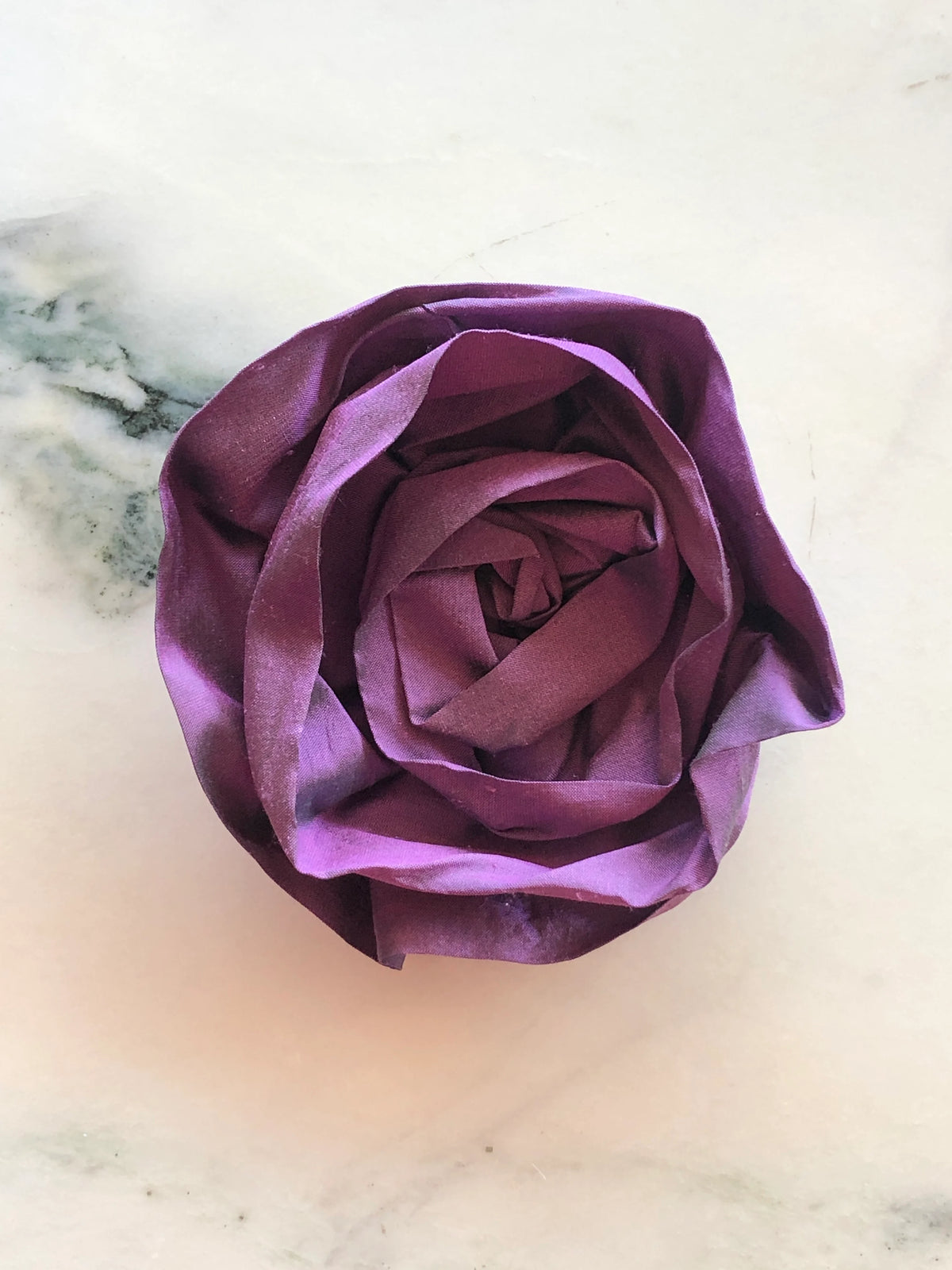 Handmade Broochella Isabella Rossellini silk flower brooch in violet, a designer statement accessory with beautifully folded rosette petals, photographed on white marble.