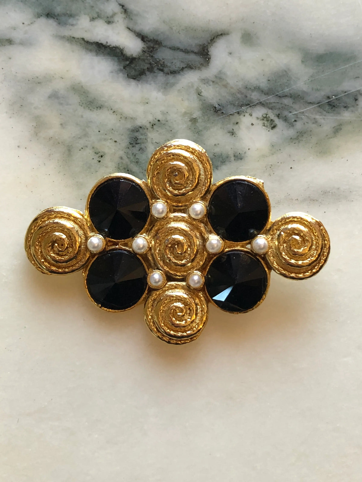 Broochella Isabella Vintage Black & Gold Statement Brooch featuring four faceted black stones, faux pearls and textured gold-tone spiral motifs on a cream and grey marble background.