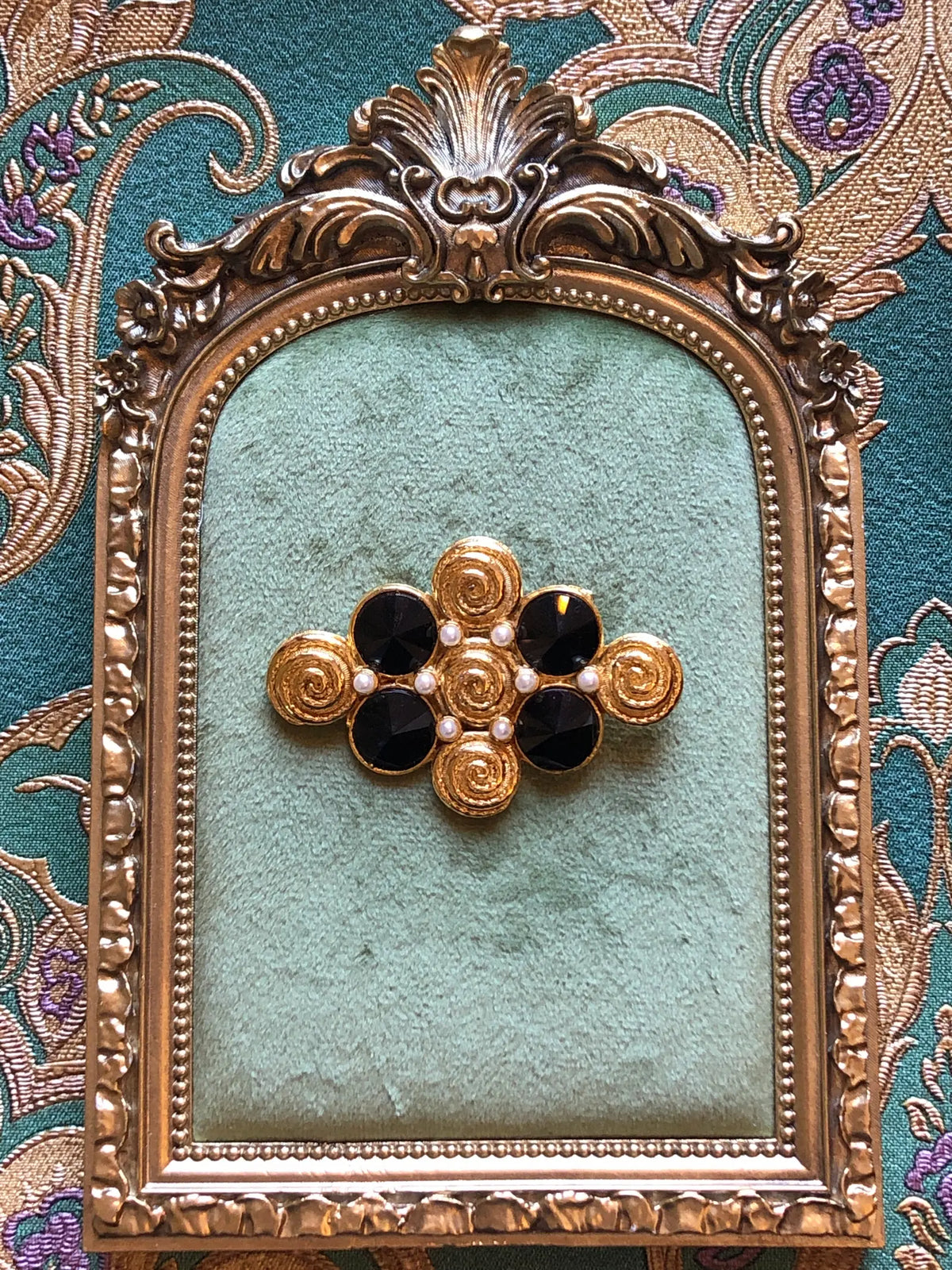 Broochella Isabella Vintage Black & Gold Statement Brooch featuring four faceted black stones, faux pearls and textured gold-tone spiral motifs. Pictured on a green velvet tray with an ornate gold frame. The background is green and old damask wallpaper.