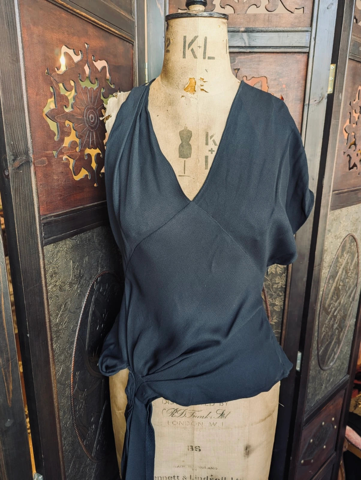 Broochella designer jacquemus le souk black multi wear draped top. It is pictured on a mannequin.The background is an ornate dark wood decorative room divider. 