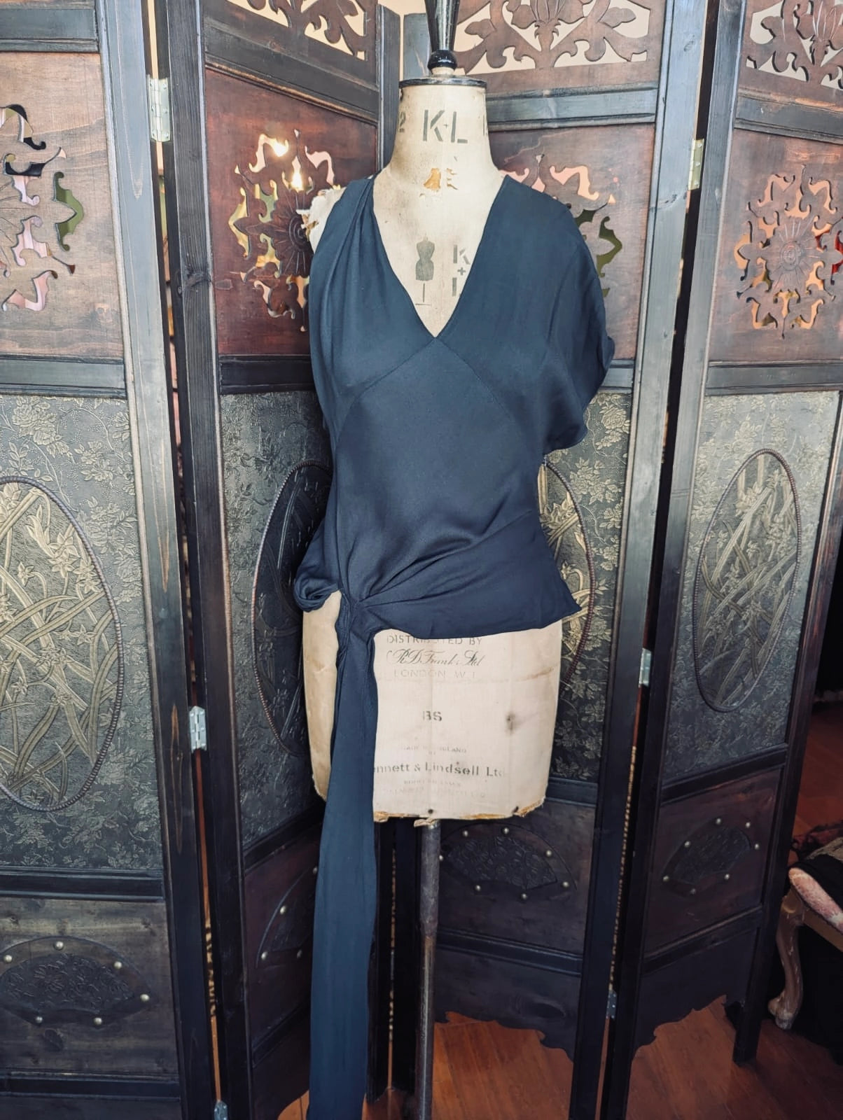 Broochella designer jacquemus le souk black multi wear draped top. It is pictured on a mannequin.The background is an ornate dark wood decorative room divider. 