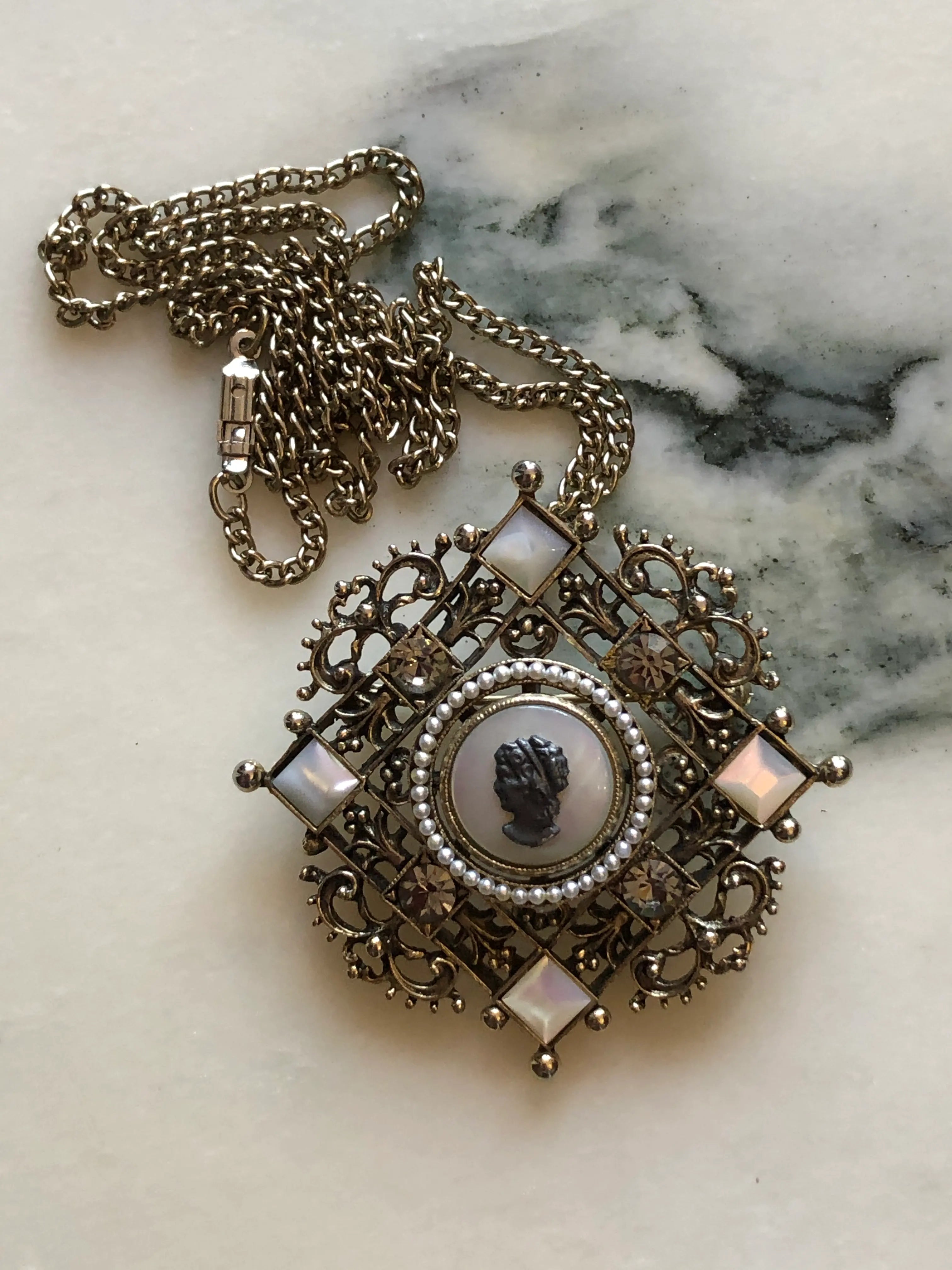 Broochella Kaiserin vintage German cameo brooch pendant necklace with ornate gold-tone filigree, seed pearls and crystal accents. It is pictured on a white marble surface