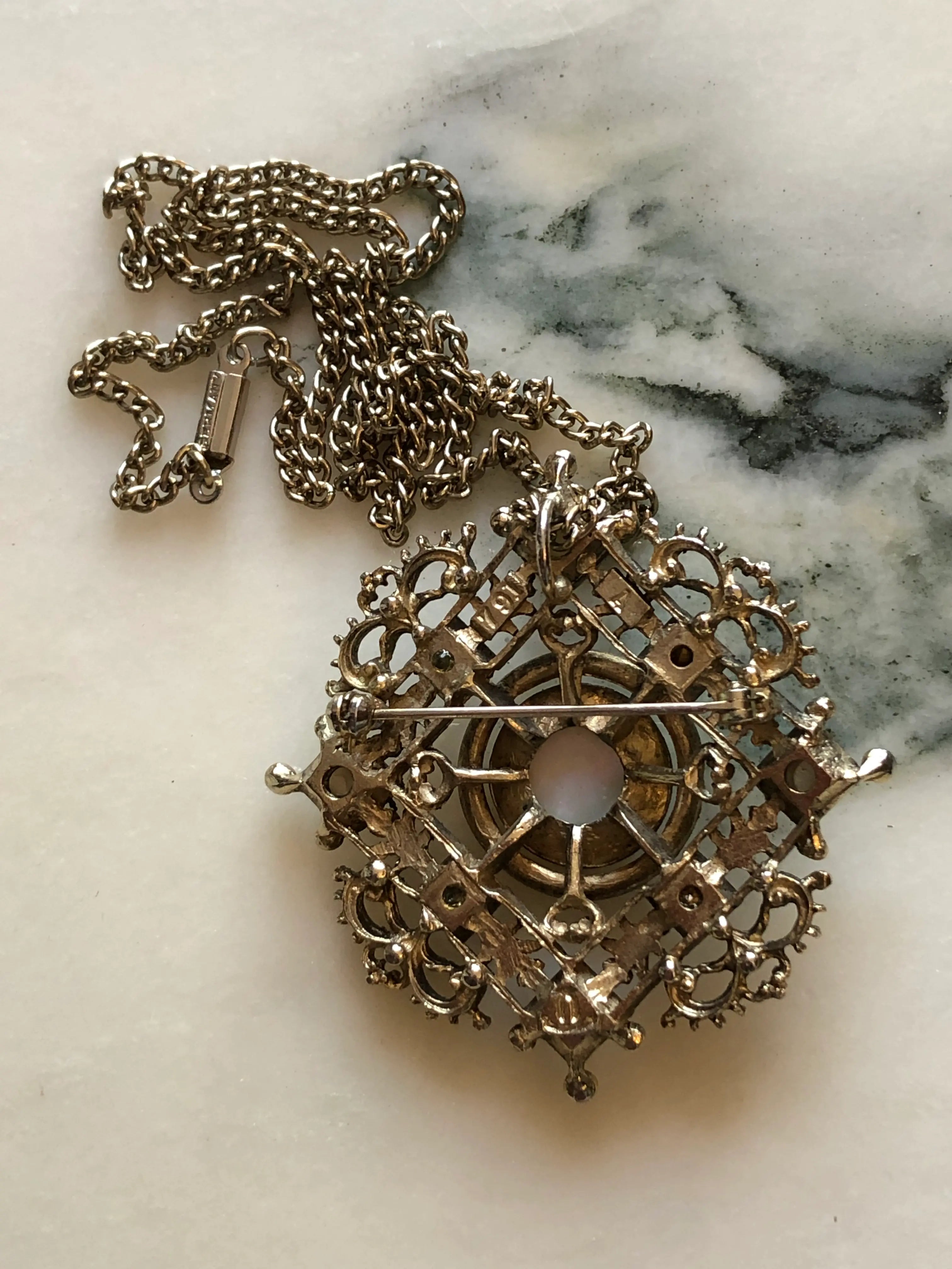 Broochella Kaiserin vintage German cameo brooch pendant necklace with ornate gold-tone filigree, seed pearls and crystal accents. It is pictured on a white marble surface