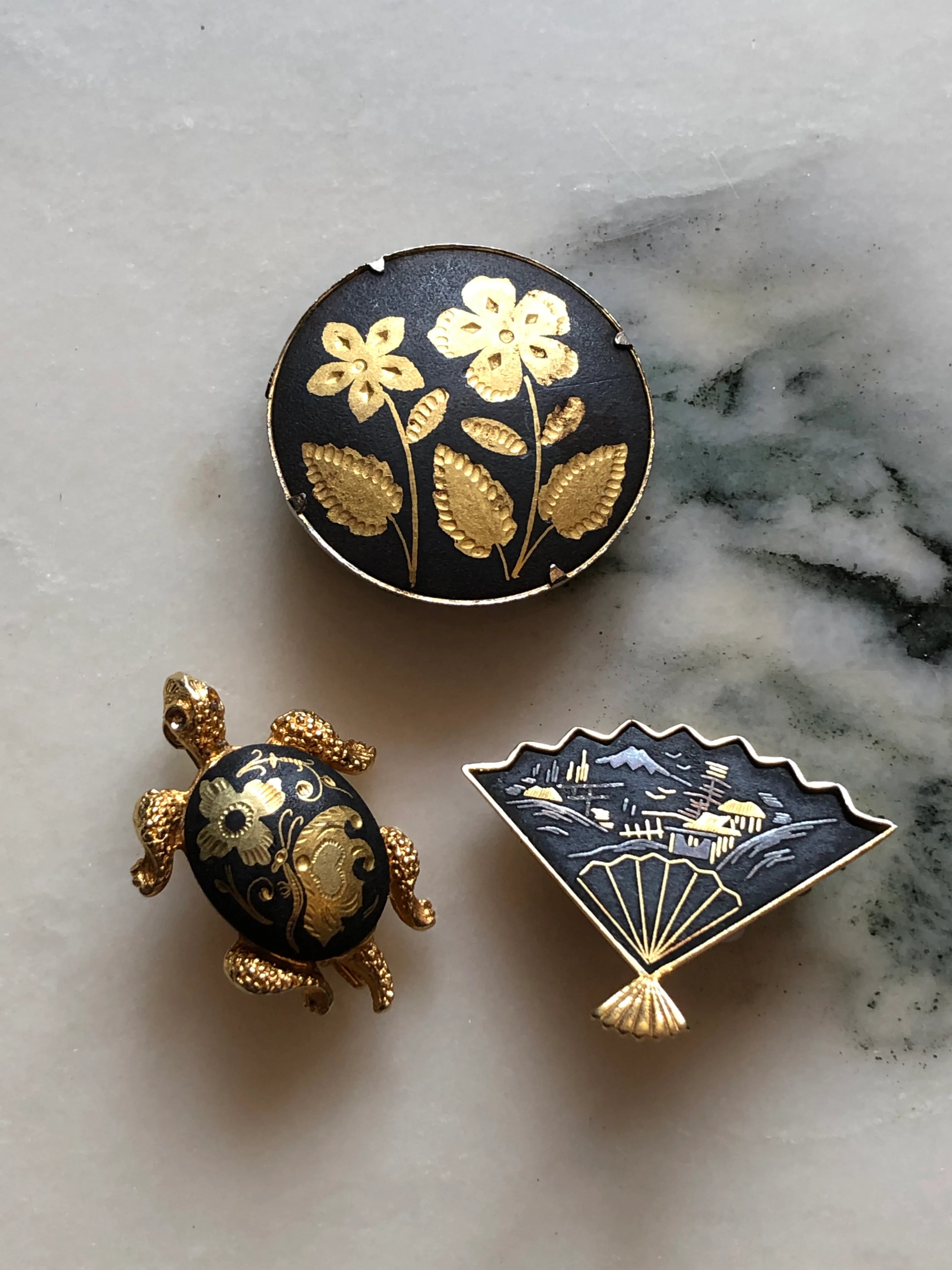 Broochella Kyoto Noir vintage black and gold brooch trio featuring an Amita Japan fan, floral medallion and decorative turtle. The background is white marble.
