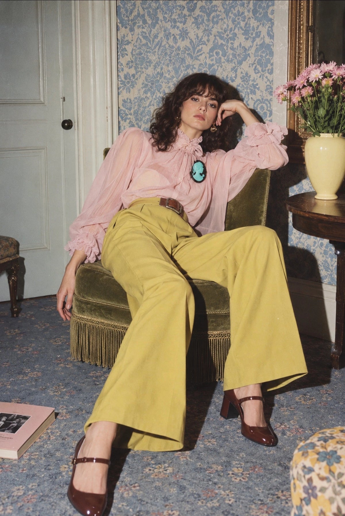 A woman sits in a vintage green armchair in a retro-style room, wearing a light pink ruffled blouse, wide-leg mustard yellow trousers, and dark brown Mary Jane heels. She is wearing the Broochella La Bella Figura Cameo Brooch in aqua.