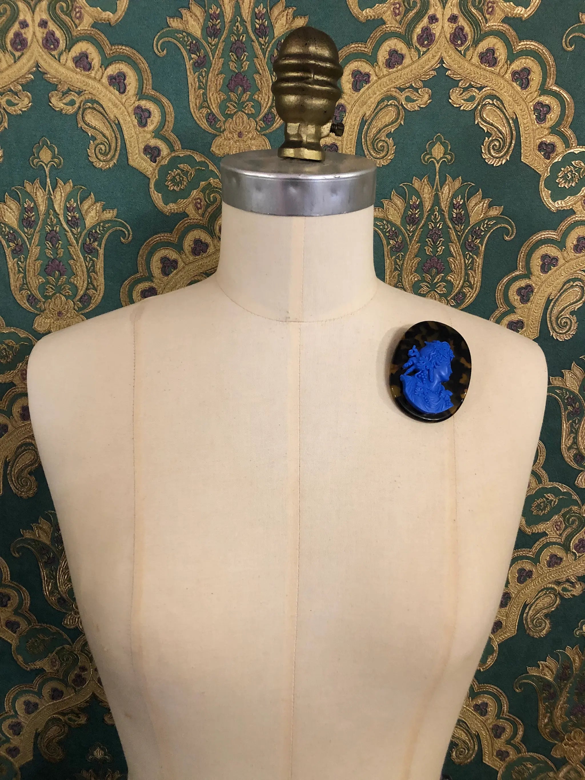 A Broochella La Bella Figura Tortoiseshell Cameo  oval brooch with a vibrant blue cameo-style face on a black background, displayed on a cream-coloured dress form against a green and gold patterned wallpaper backdrop.