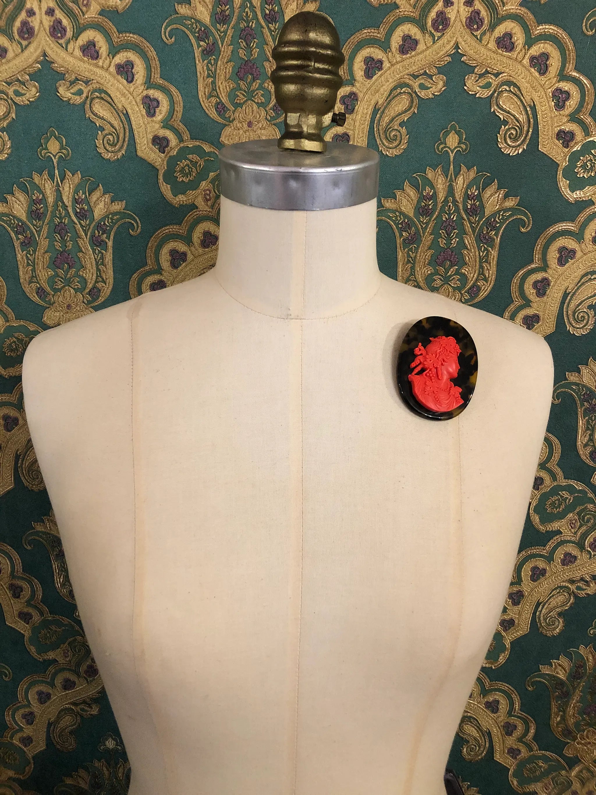 Broochella La Bella Figura Cameo brooch. A red cameo on a tortoiseshell base,  displayed on a cream dress form against a dark teal and gold patterned fabric backdrop.