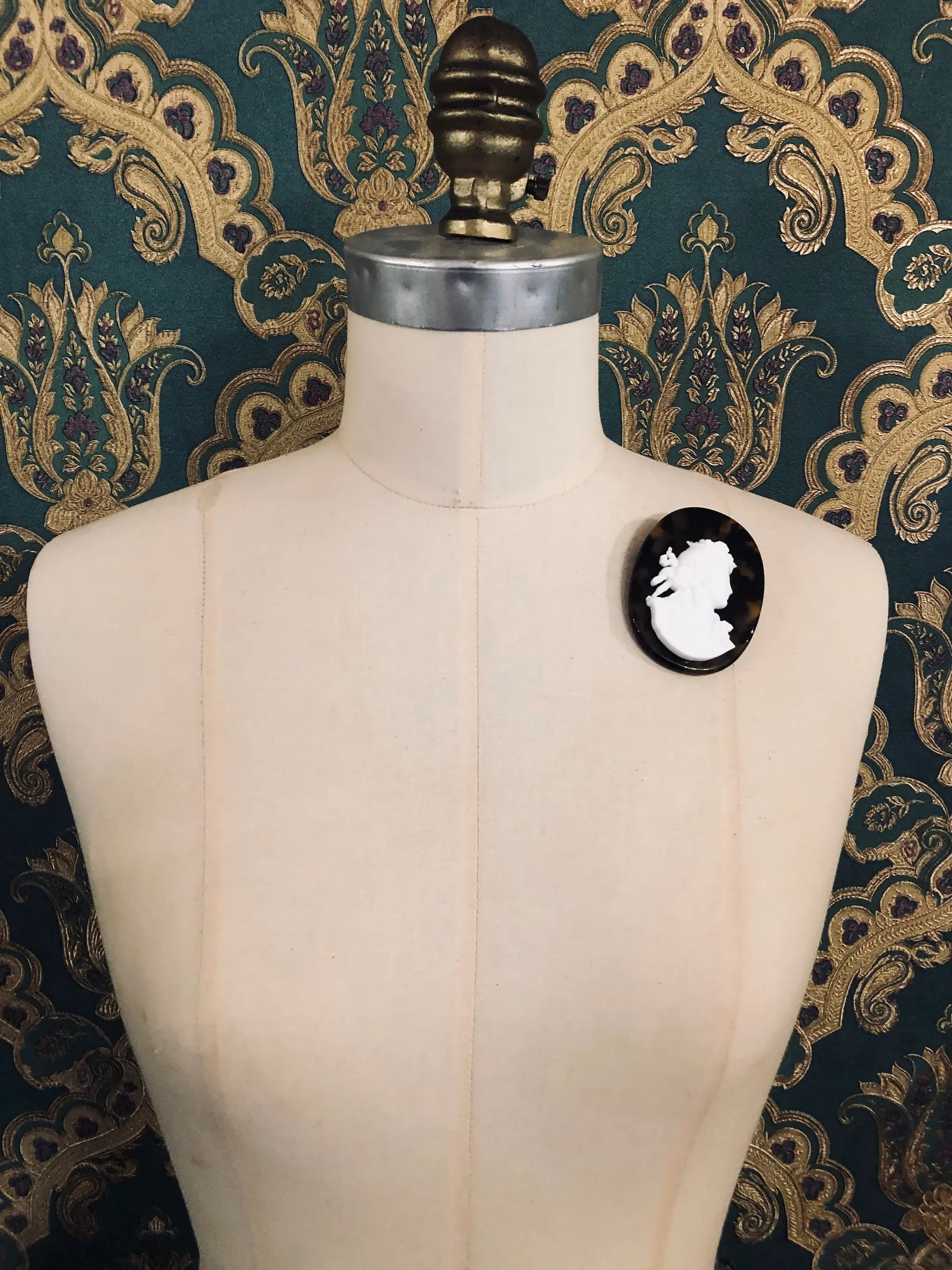 Broochella La Bella Figura Cameo brooch. A White cameo on a tortoiseshell base,  displayed on a cream dress form against a dark teal and gold patterned fabric backdrop.