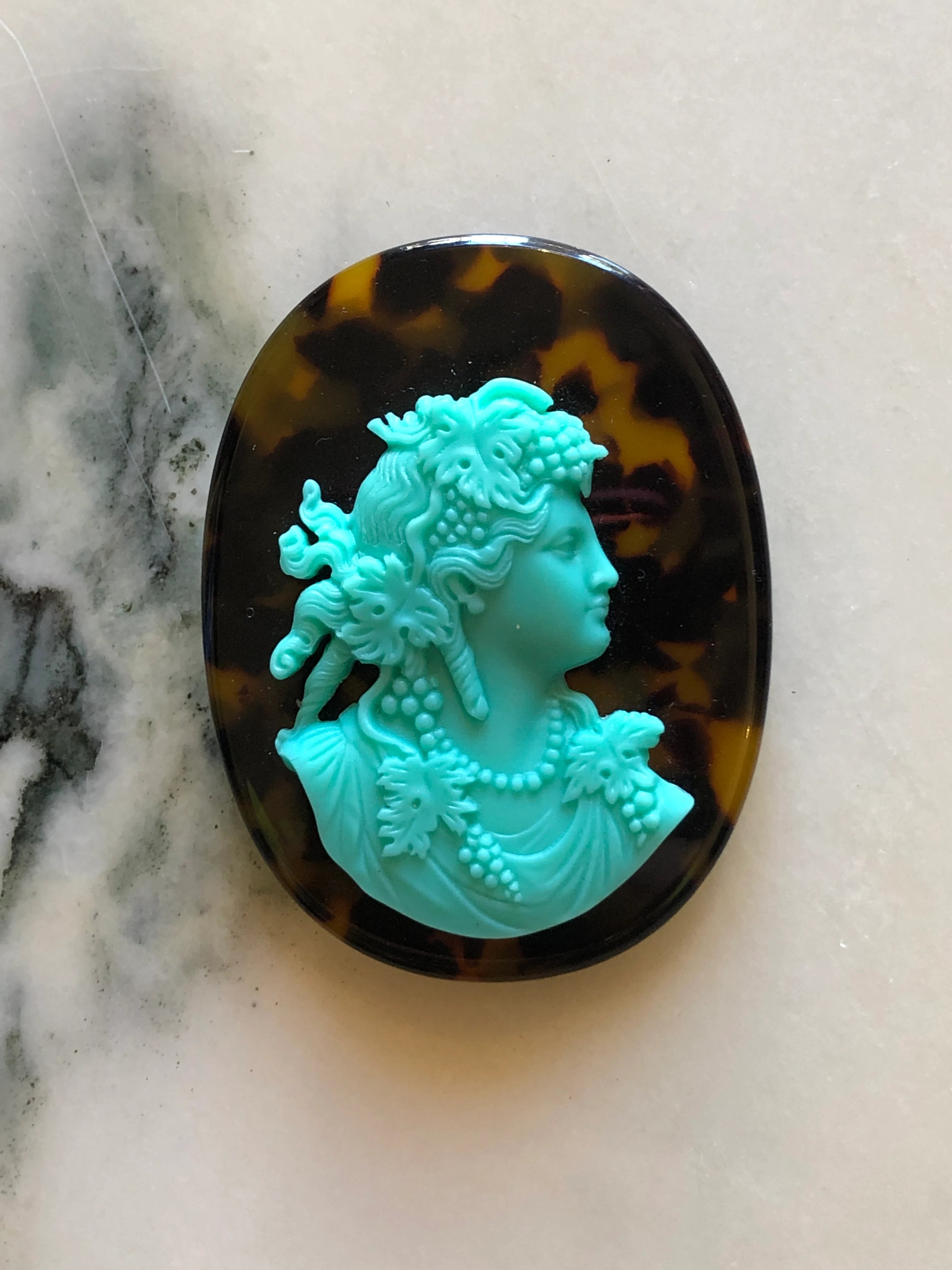 A Broochella La Bella Figura oval cameo brooch featuring an aqua classical profile bust of a woman with floral details, set against a dark brown and amber tortoiseshell-patterned background, displayed on a light marble surface.