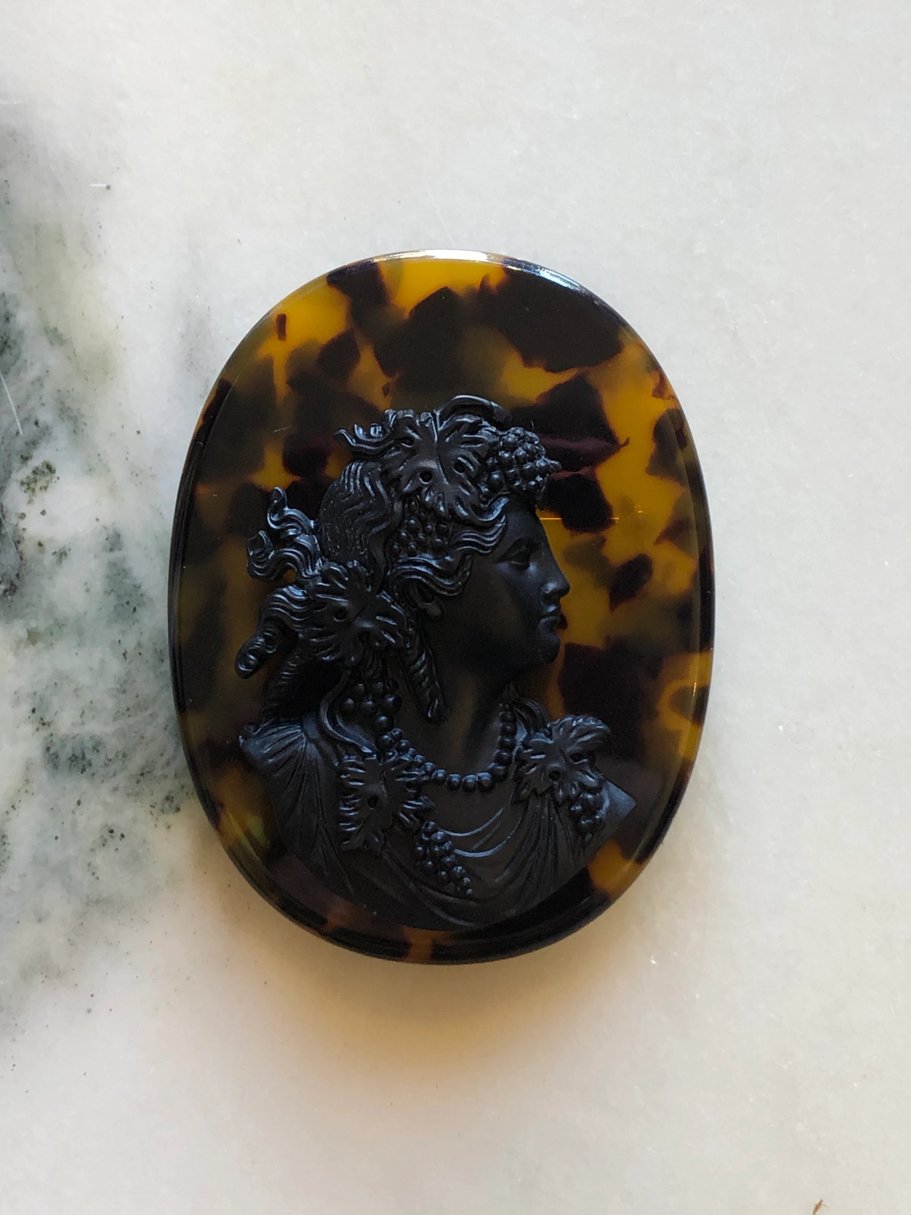 A Broochella La Bella Figura oval cameo brooch featuring a black classical profile bust of a woman with floral details, set against a dark brown and amber tortoiseshell-patterned background, displayed on a light marble surface.