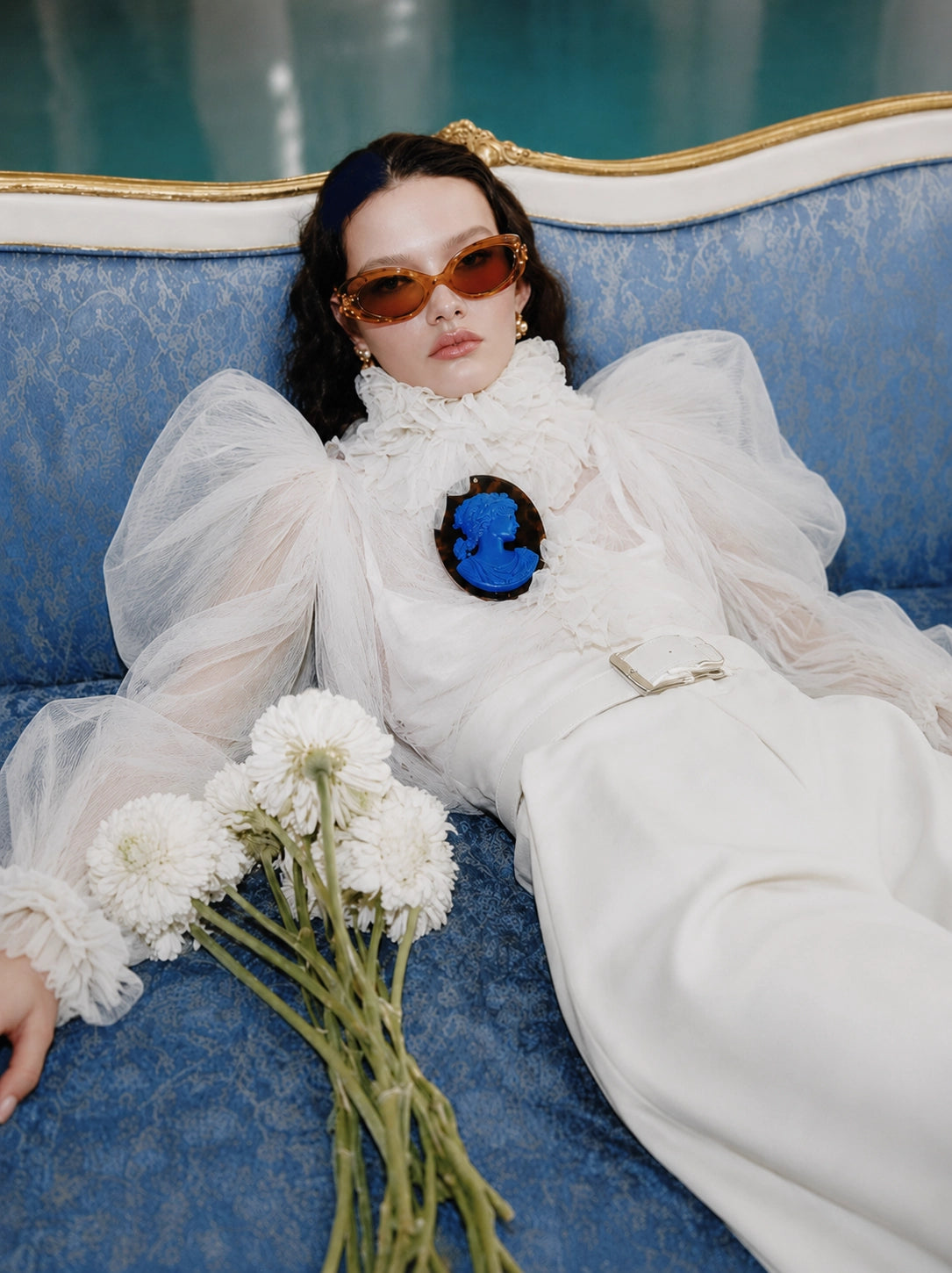 A model reclining on a blue patterned sofa wearing a white ruffled tulle dress, orange-tinted sunglasses, and a Broochella Bella Figura blue tortoiseshell cameo brooch, with white flowers in the foreground.