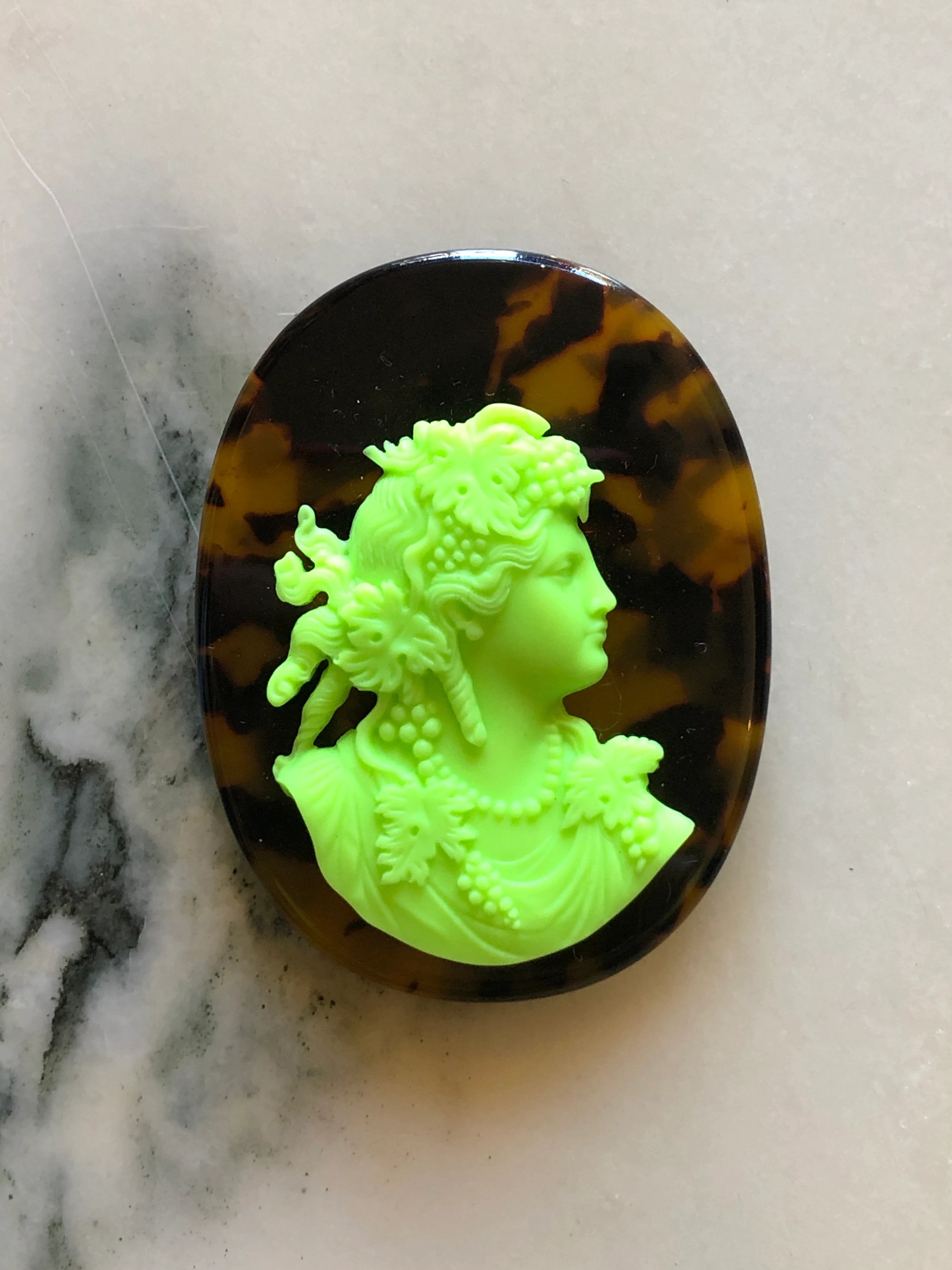 A Broochella La Bella Figura Tortoieshell oval cameo pendant or brooch featuring a bright neon green classical profile bust of a woman with floral details, set against a dark brown and amber tortoiseshell-patterned background, displayed on a light marble surface.