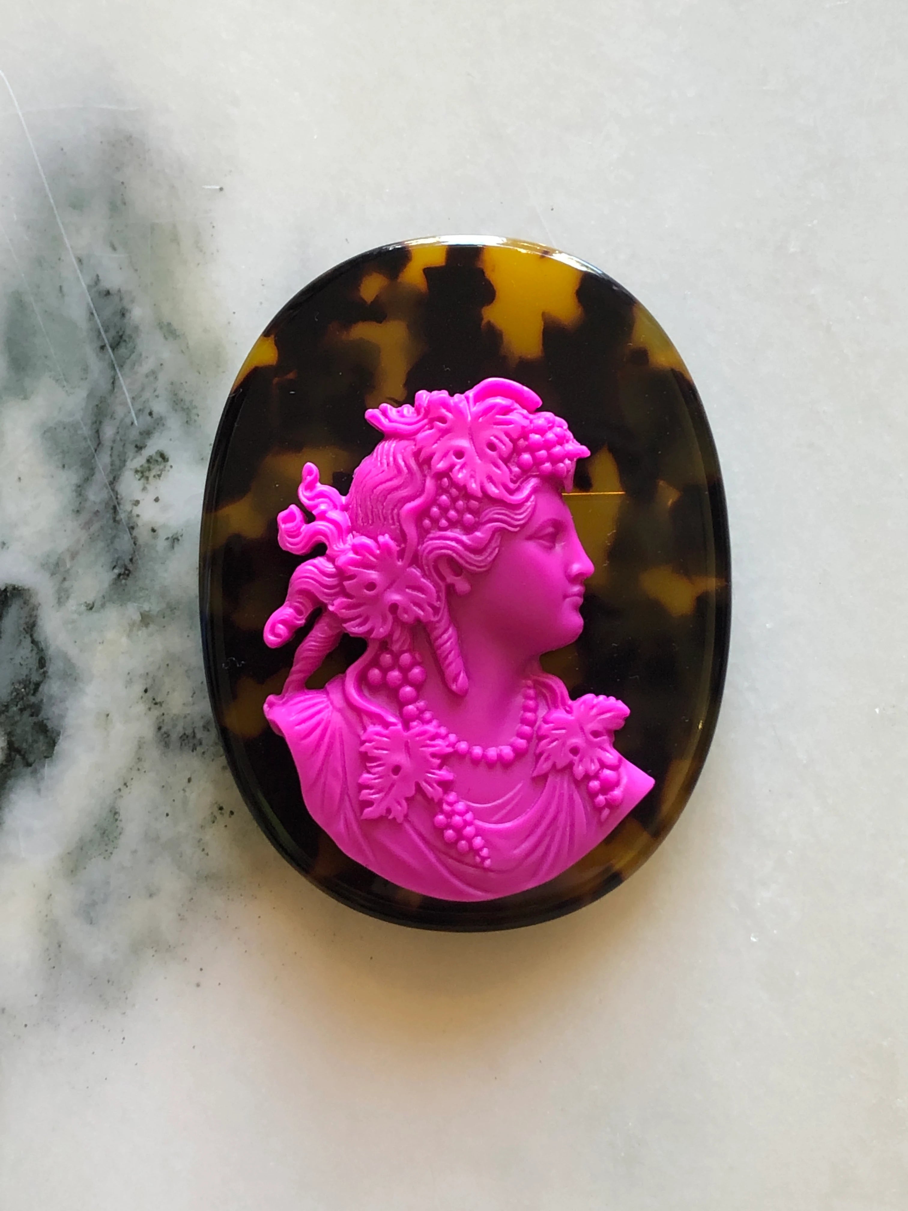 An oval Broochella La Bella Figura Tortoiseshell cameo brooch featuring a bright pink classical bust profile in the centre, set against a dark brown and black tortoiseshell background, displayed on a light marble surface.