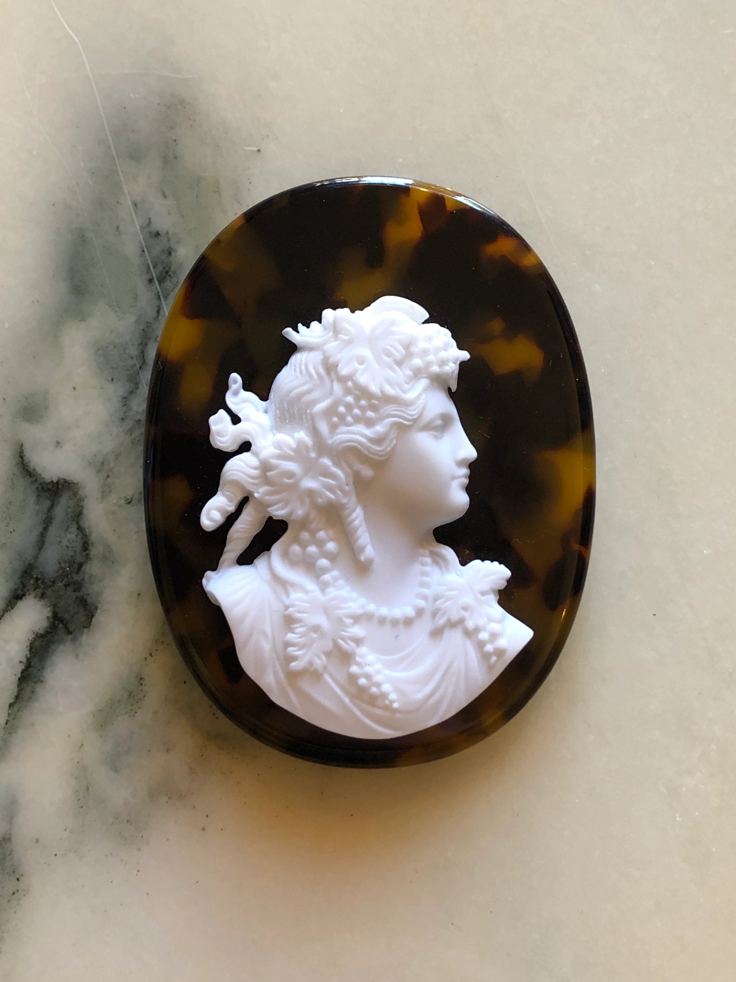 An oval Broochella La Bella Figura Tortoiseshell cameo brooch featuring a white classical bust relief set in a dark tortoiseshell frame, photographed on a light marble-like surface.