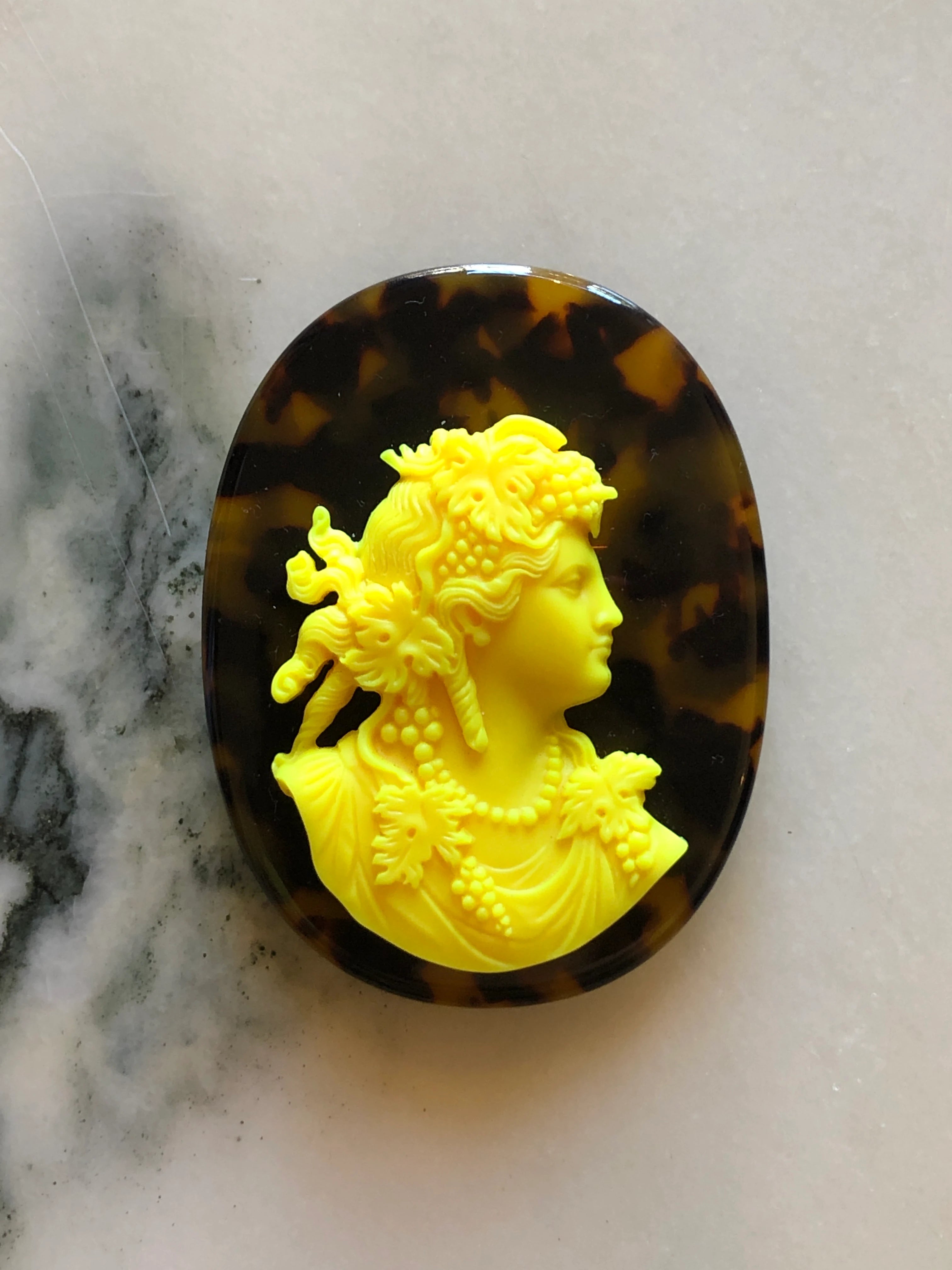 An oval Broochella La Bella Figura Tortoiseshell cameo brooch featuring a bright yellow classical bust relief of a woman in profile, set against a dark brown and black tortoiseshell background, displayed on a light marble surface.