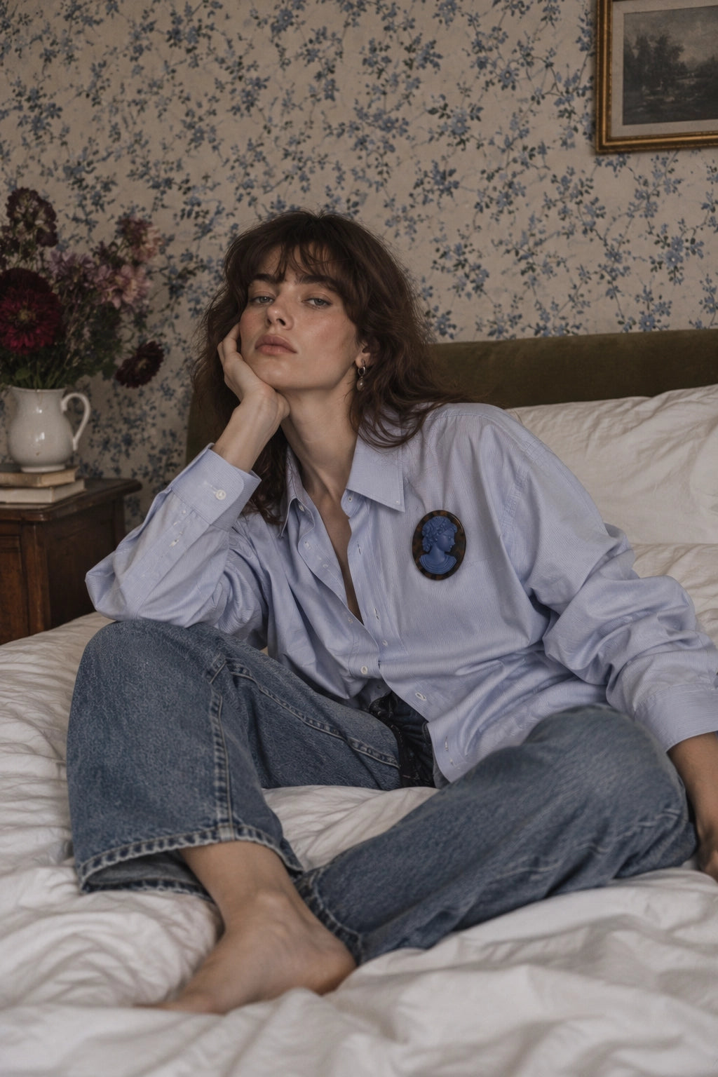 A woman sits on a bed wearing a light blue button-up shirt with a Broochella blue La Bella Figura tortoiseshell cameo brooch on the chest and blue jeans, set against a background of floral wallpaper and a wooden headboard.