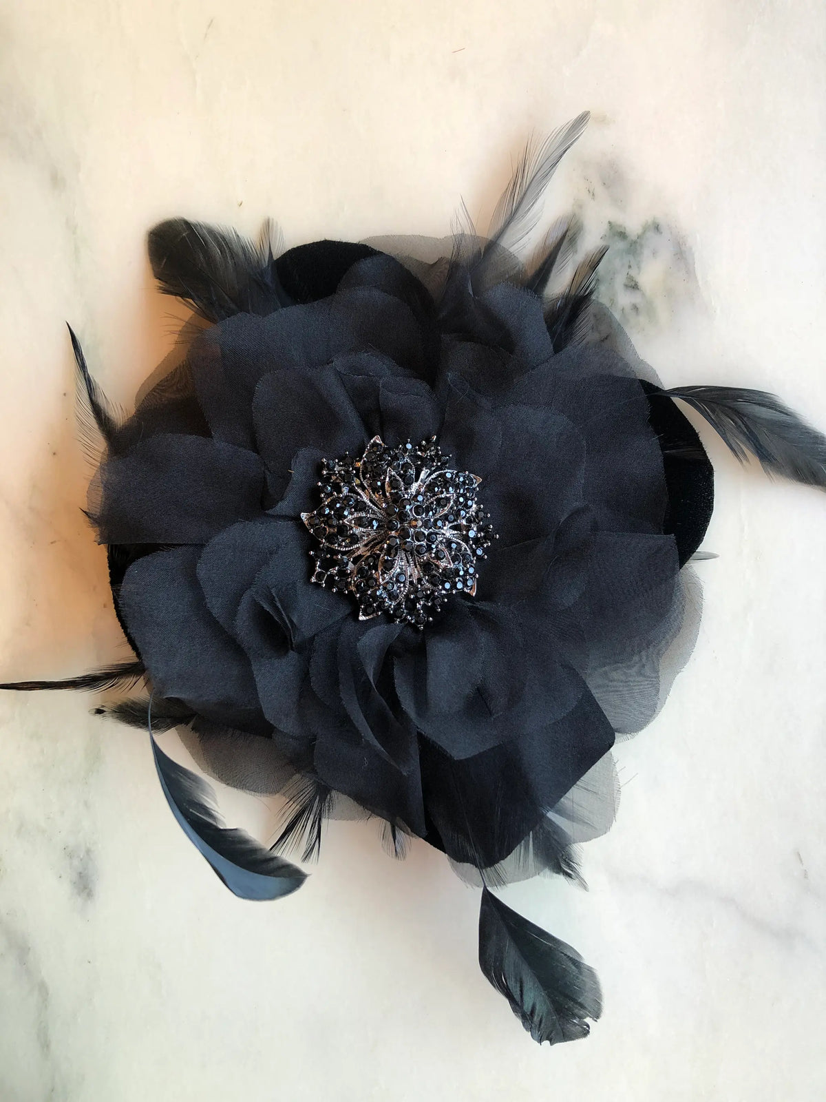 Broochella La Contessa Flower Brooch Pin in black, featuring layered fabric petals, delicate black feathers and an ornate black crystal centre, photographed on a white marble background with soft grey veining.