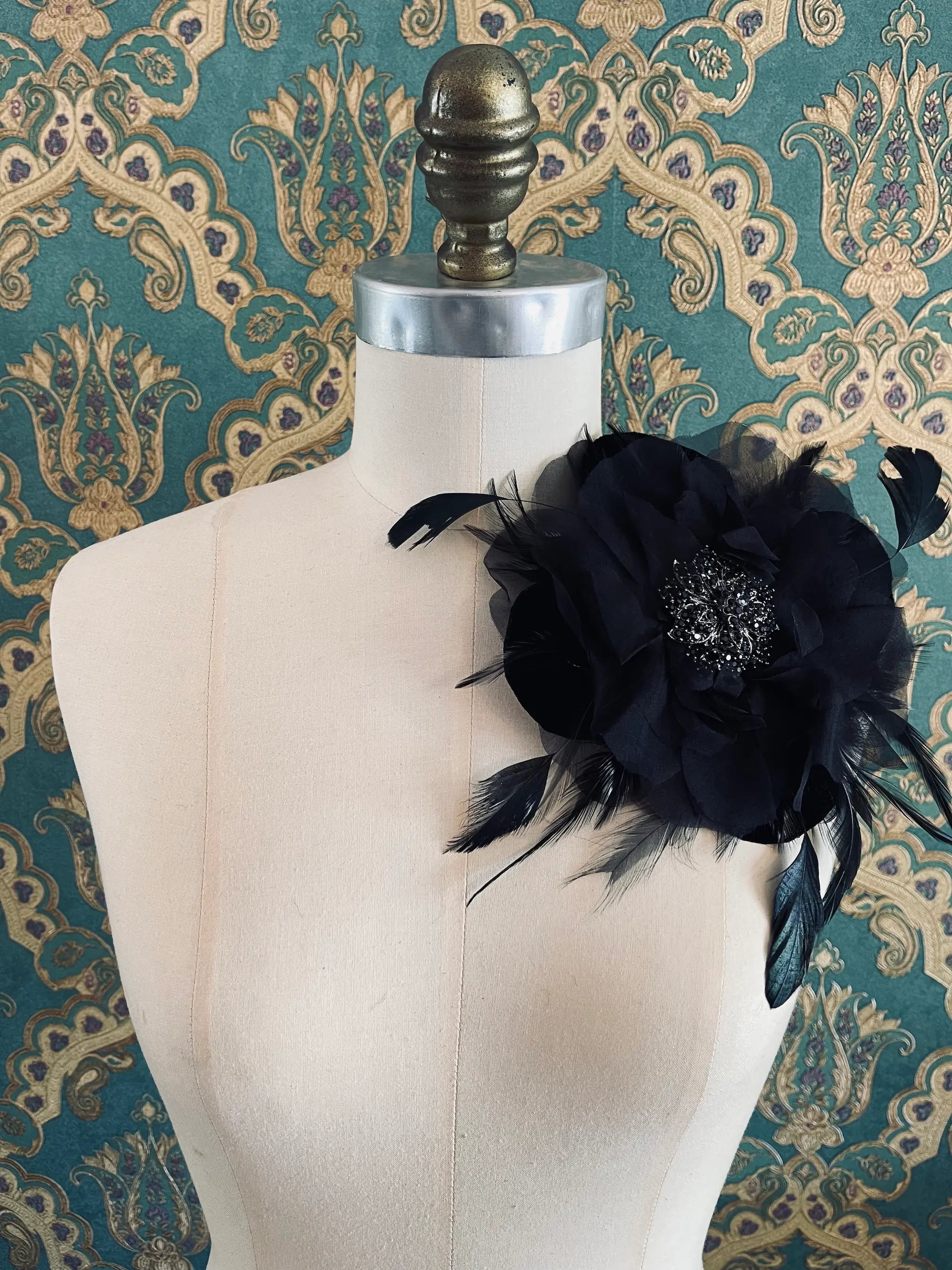 Broochella la contessa handmade fabric and feather flower brooch with centre embellishment. Pictured on a mannequin. The background is green and gold damask wallpaper