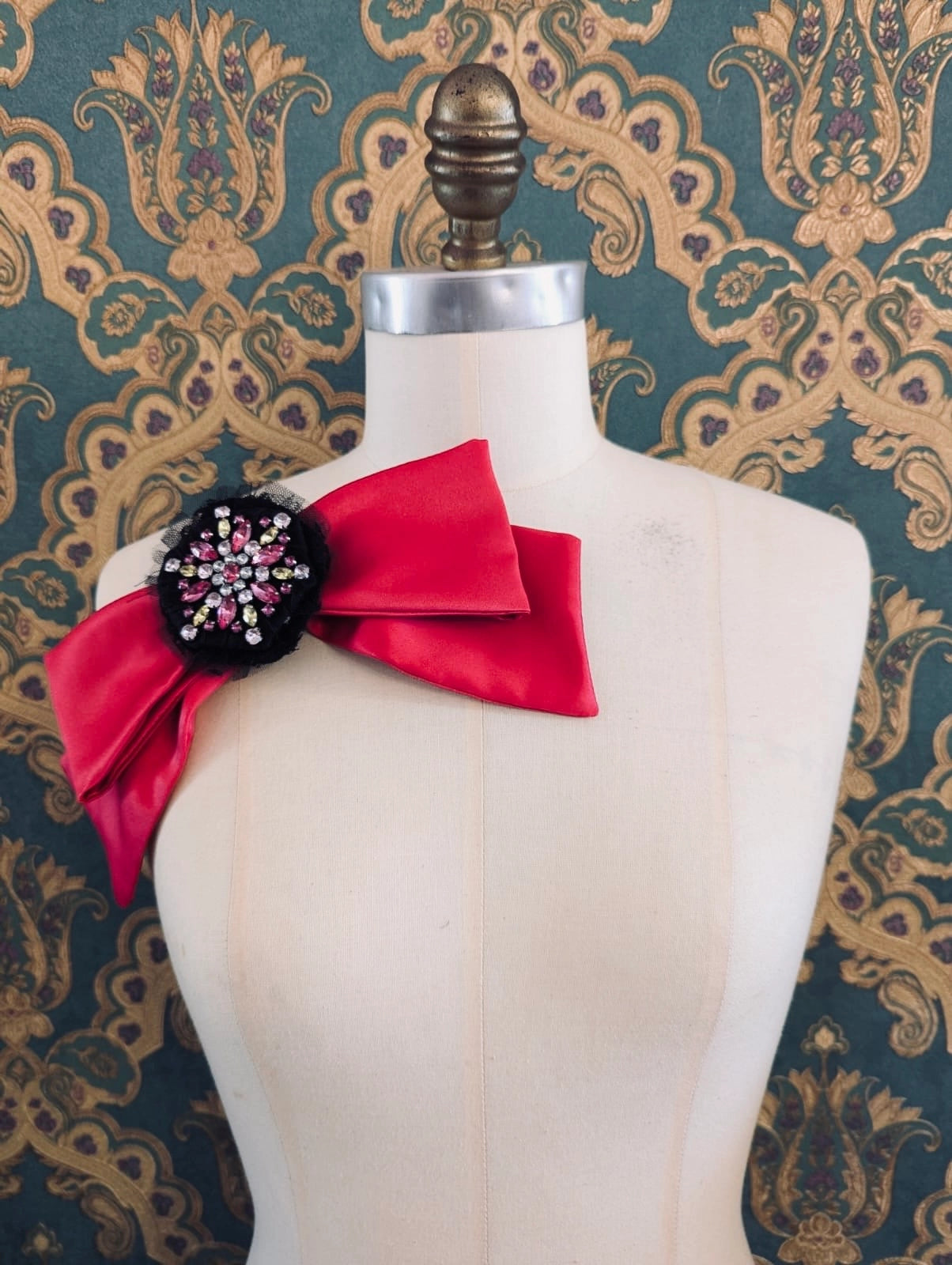 Broochella la dolce vita handmade pink satin bow. It had a black and pink brooch embellishment at the centre if the bow.  It is pinned to a mannequin. The background is green and gold damask wallpaper.