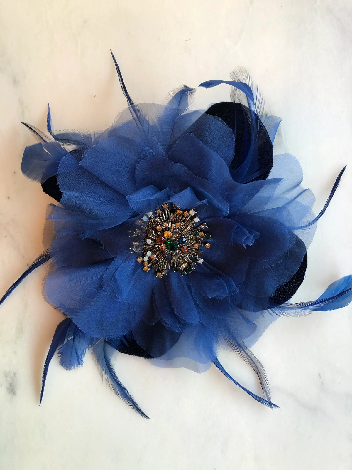 Broochella Men's La Duchessa Flower Brooch Pin in deep royal blue, featuring layered sheer fabric petals, sweeping blue feathers and an ornate multicoloured jewelled centre, photographed on a white marble background with soft grey veining.