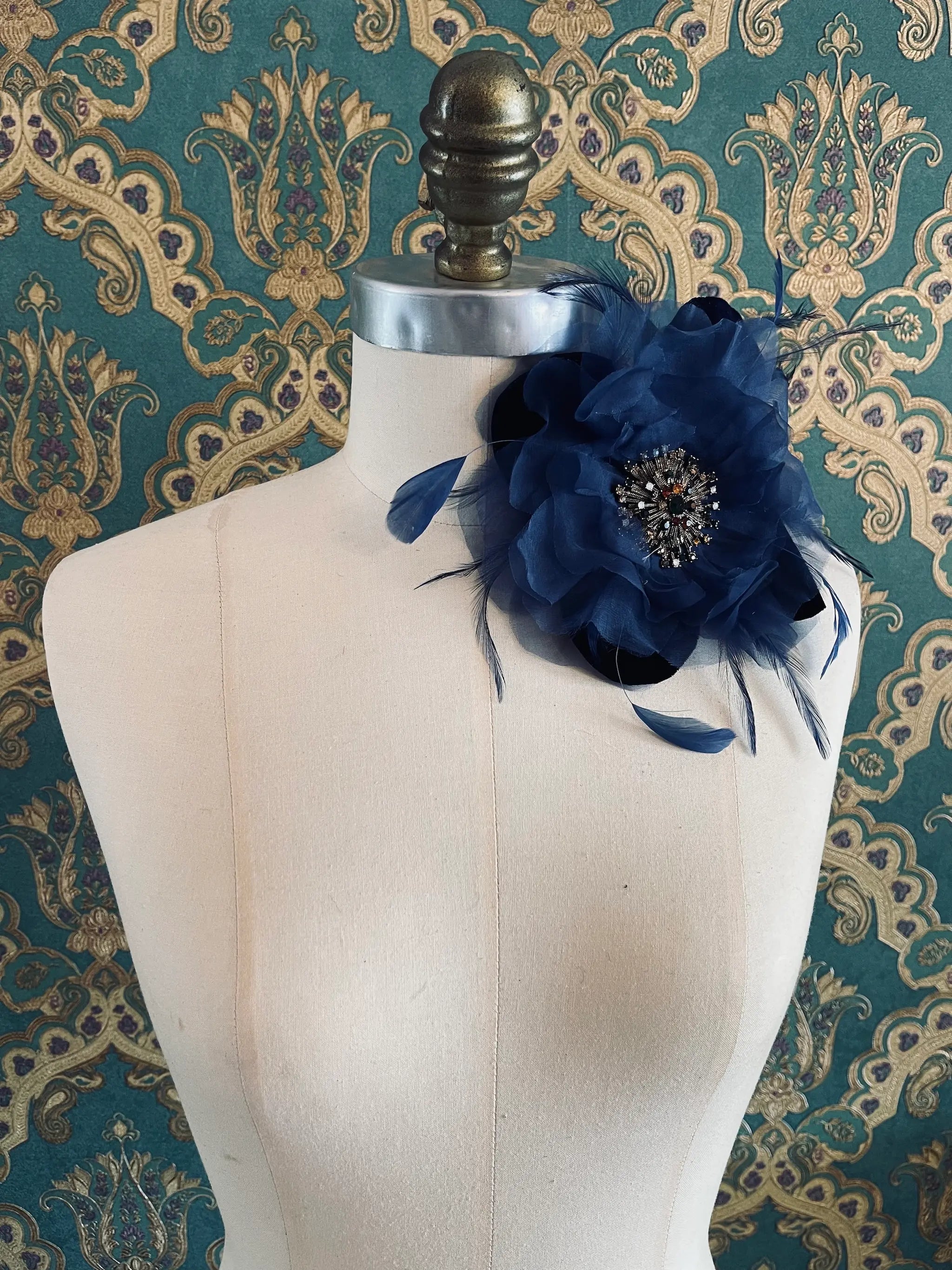 Broochella la duchessa blue handmade fabric and feather flower brooch with centre embellishment. Pictured on a mannequin. The background is green and gold damask wallpaper