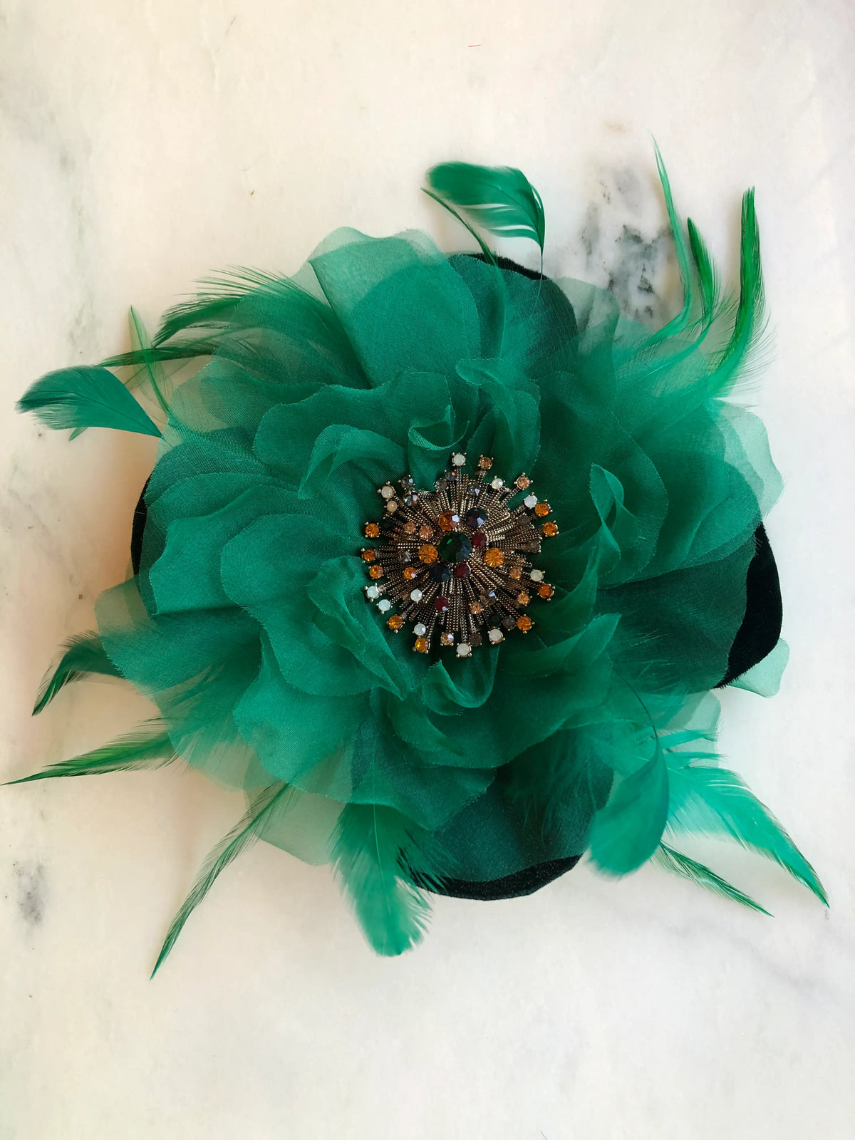 Broochella La Duchessa Flower Brooch Pin in emerald green, featuring voluminous sheer fabric petals, dramatic green feathers and an ornate multicoloured jewelled centre, photographed on a white marble background with soft grey veining.