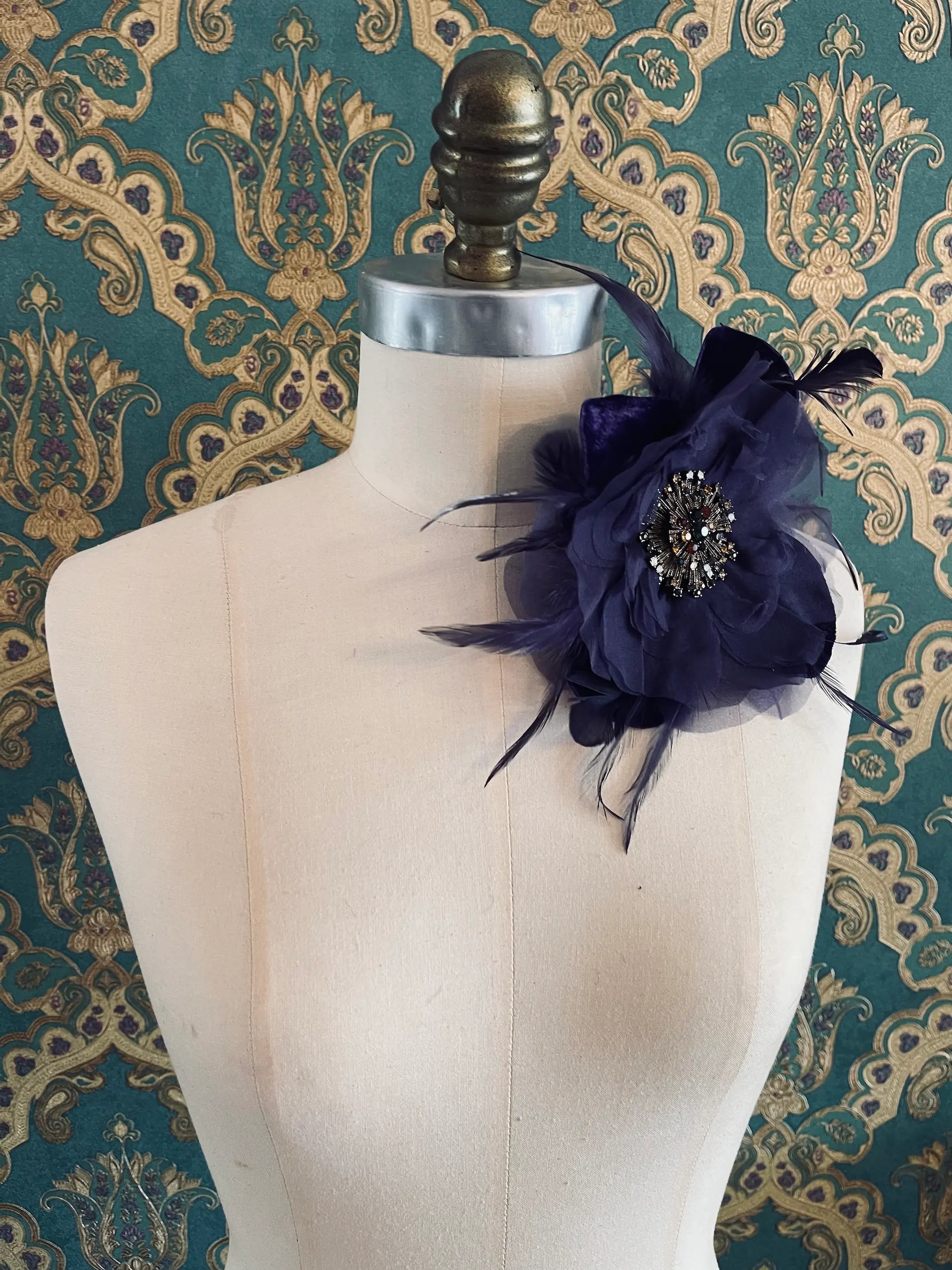 Broochella la duchessa purple handmade fabric and feather flower brooch with centre embellishment. Pictured on a mannequin. The background is green and gold damask wallpaper