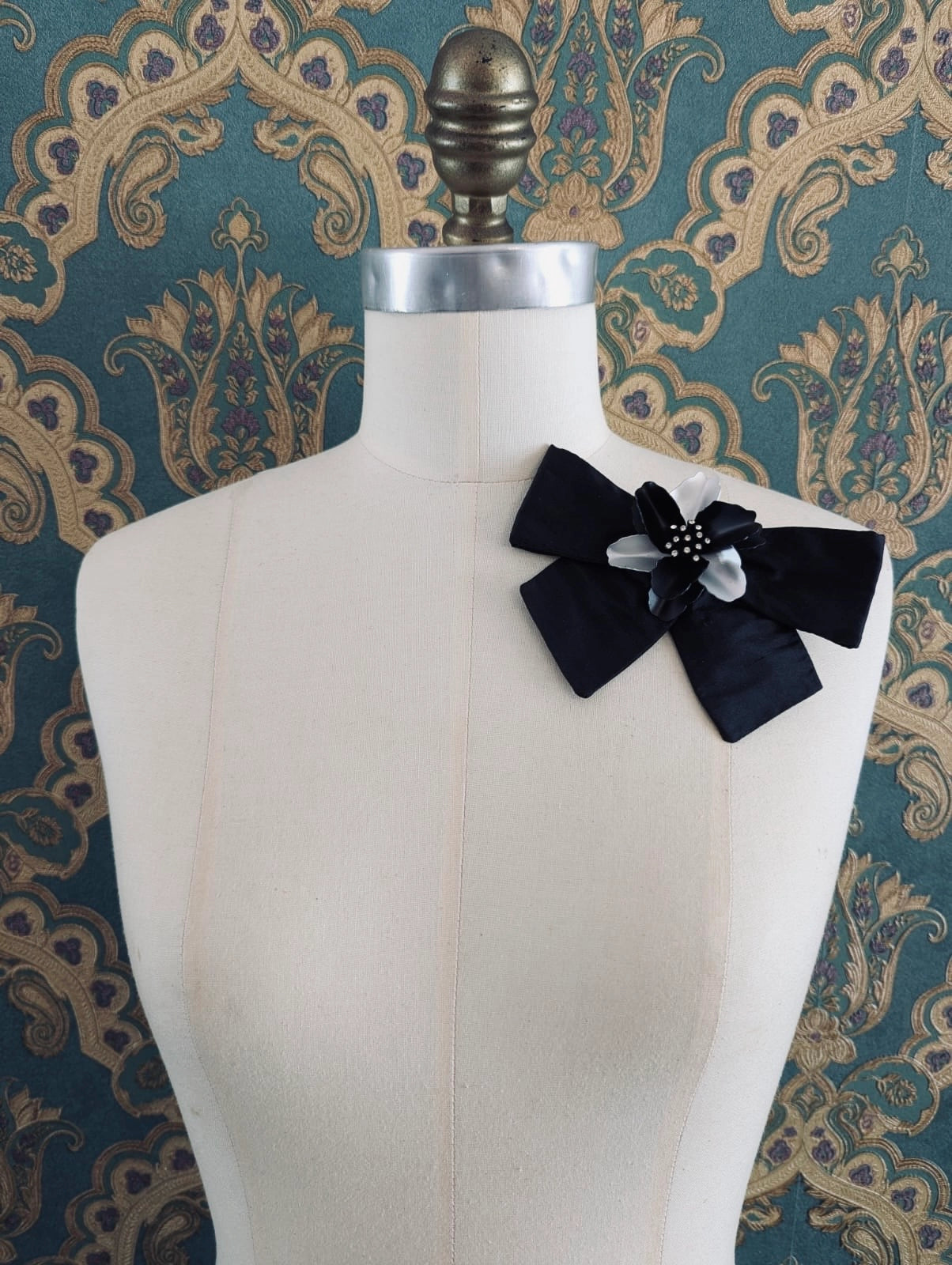 Broochella la notte handmade black silk bow brooch, with black and silver vintage flower brooch at the centre. Pinned on a mannequin. The background is green and gold damask wallpaper. 