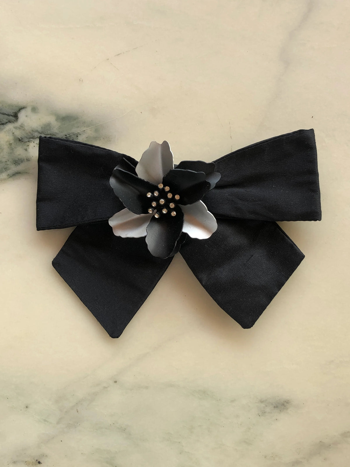 Broochella La Notte Bloom Bow Brooch in black silk featuring a sculptural black and white flower with crystal embellishments at the centre, photographed on a pale white marble background with grey veining.