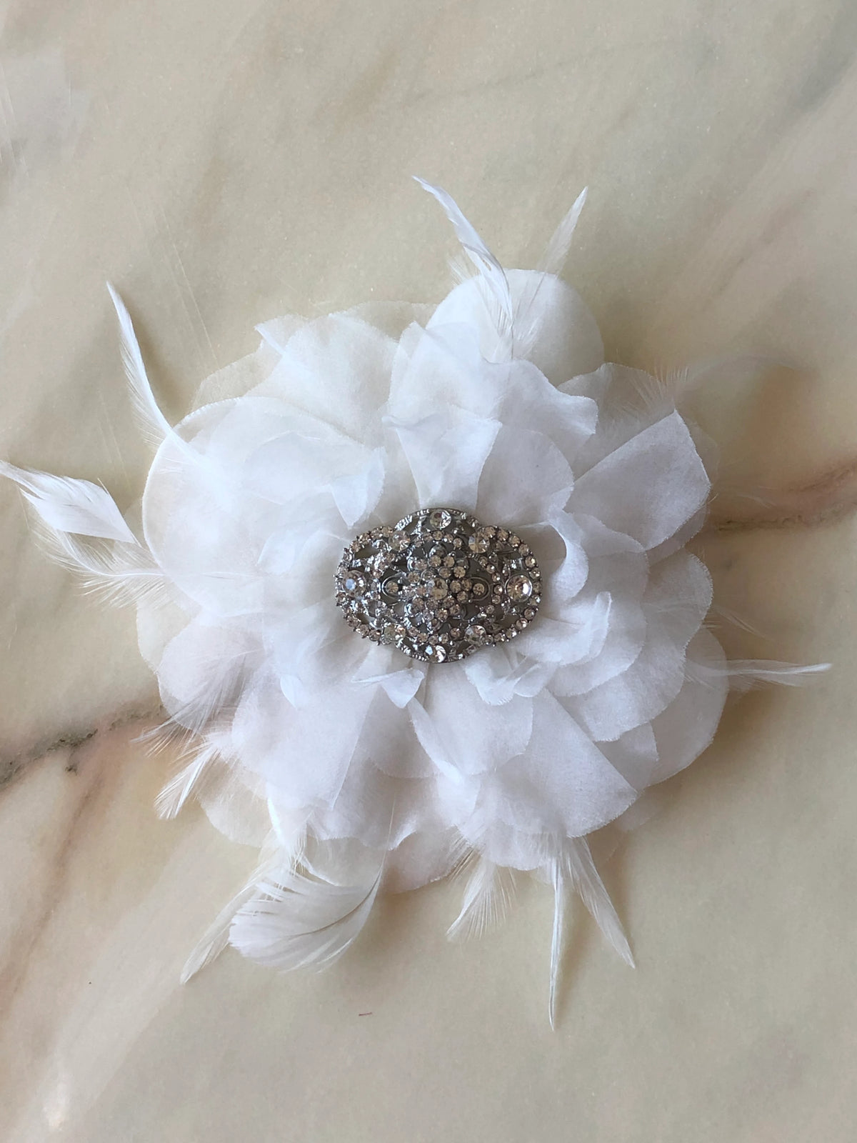 Broochella La Sposa Bridal Flower Brooch in white with delicate feathers and a crystal embellished centre, photographed on a white marble background.