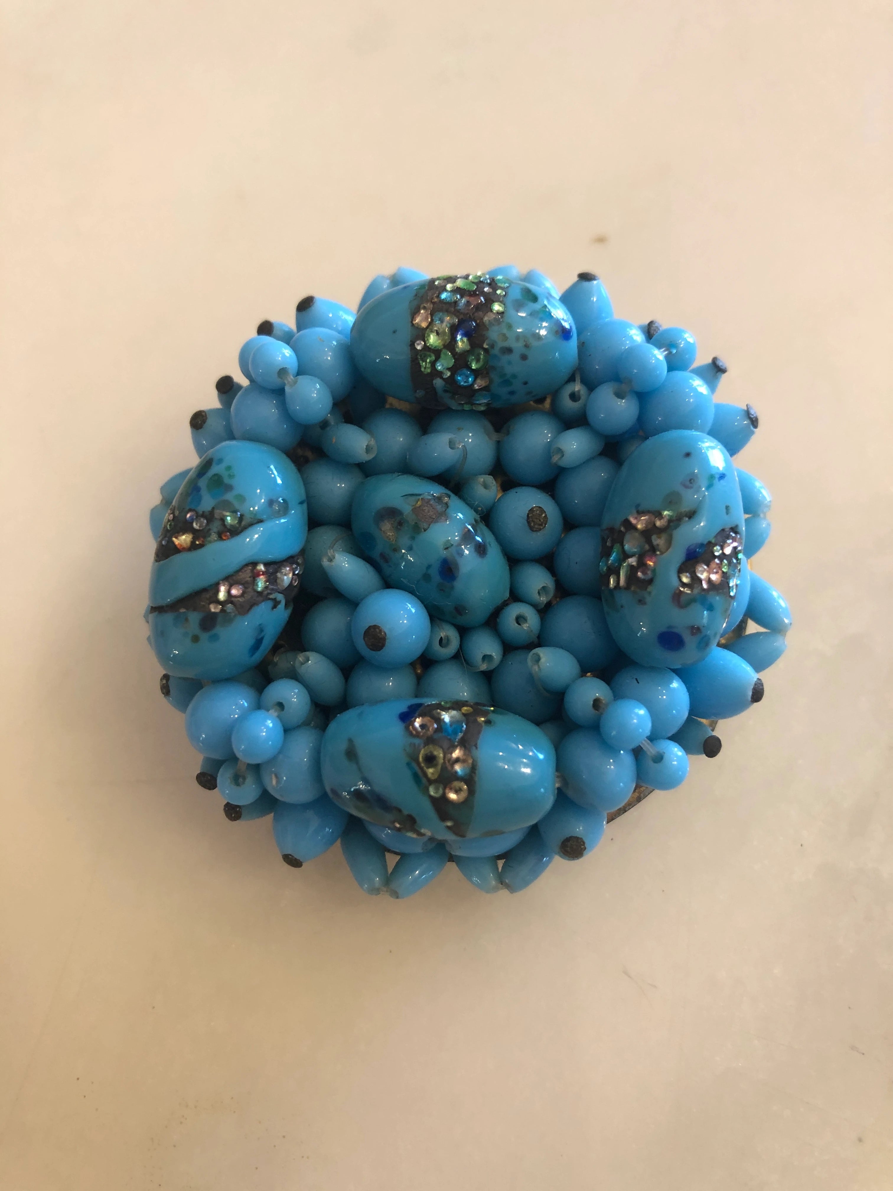 A Broochella laguna Turchese brooch with circular cluster of bright turquoise blue beads with small silver-toned floral accents. Pictured on a white marble background. 