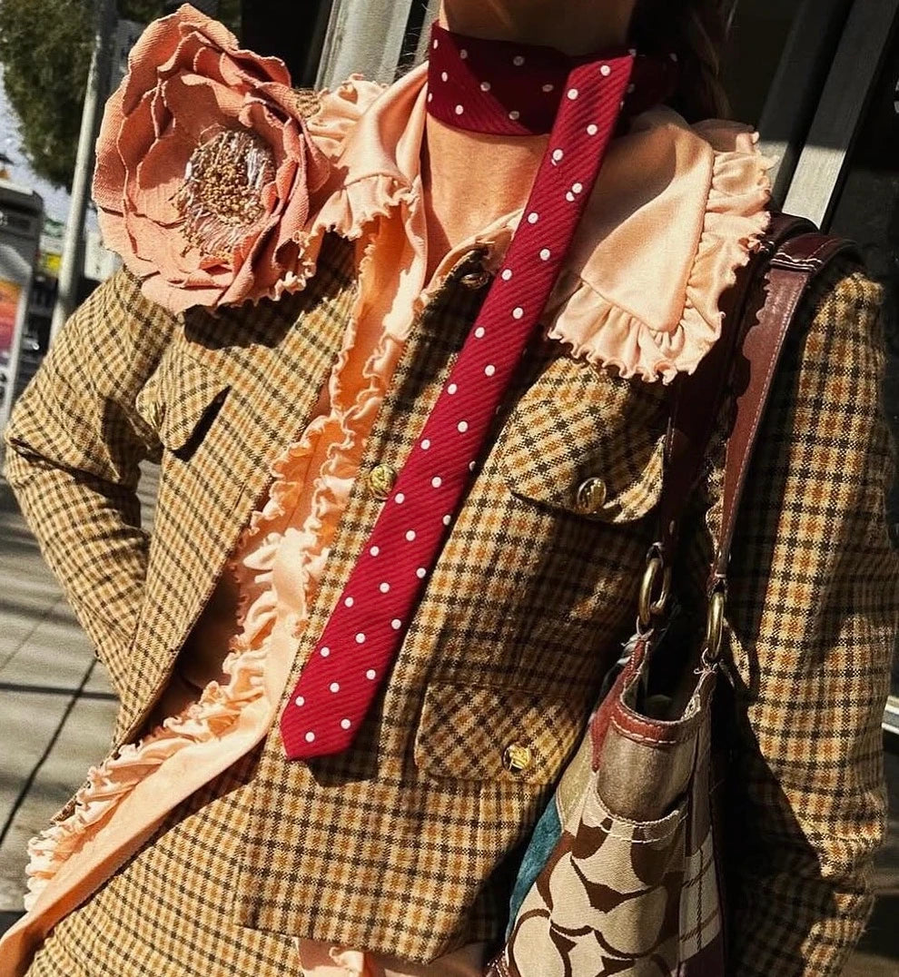 A close-up of a person wearing a brown and beige plaid blazer over a peach ruffled blouse, accessorised with a red polka-dot necktie, a large Broochella handmade Anemone  fabric flower brooch, and a patterned shoulder bag.